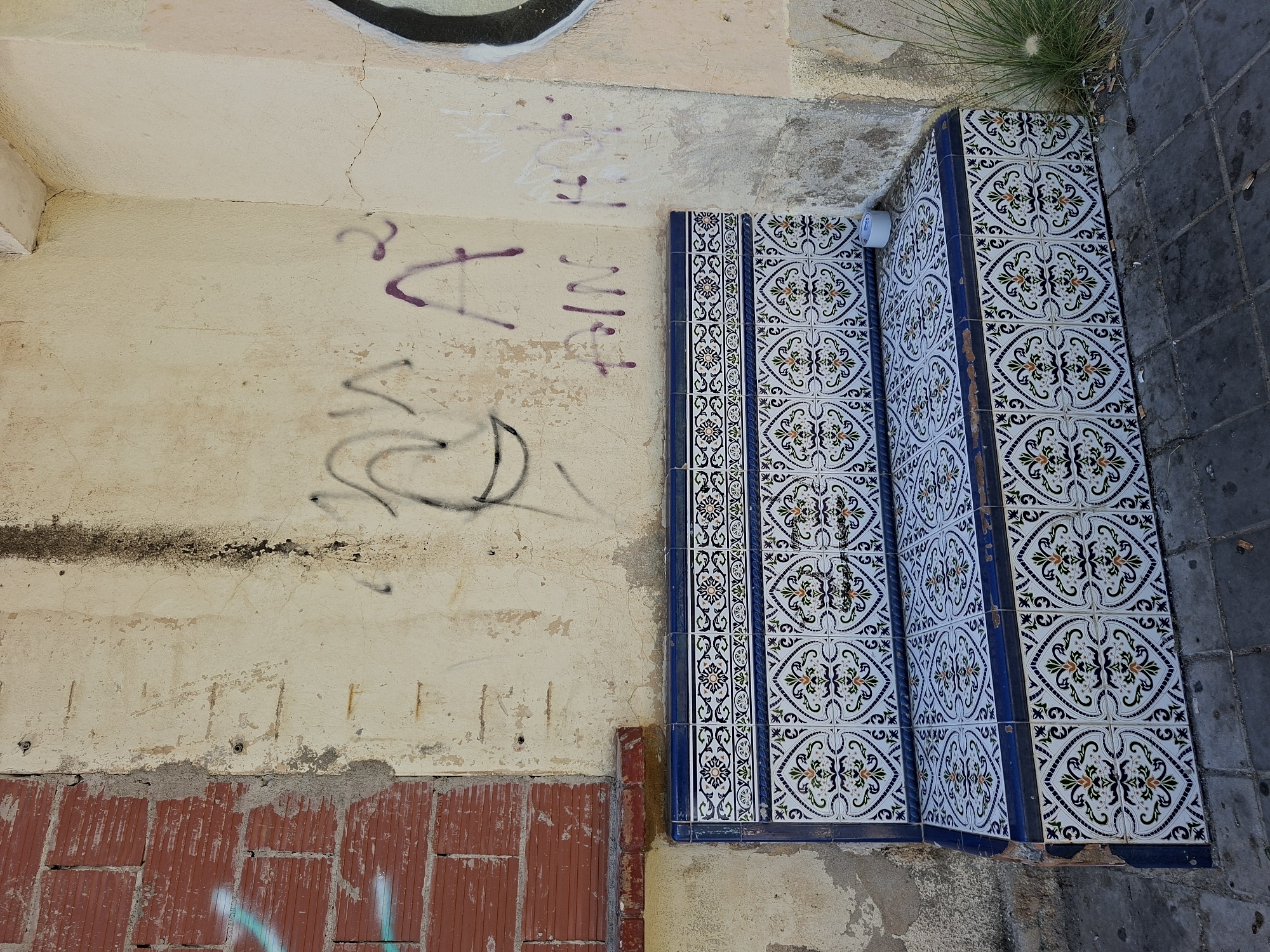 This image depicts an outdoor scene featuring a tiled structure, likely part of a staircase or seating area, adjacent to a weathered wall with graffiti. The tiles are ornate, featuring a blue border and a white background with intricate floral patterns in black, green, and orange. The wall is painted a pale yellow and shows signs of age, with cracks and peeling paint. Graffiti in purple and black spray paint is visible on the wall. Below the yellow wall, a section of red brickwork is exposed, also bearing graffiti. The ground next to the tiled structure appears to be paved with dark grey stones. A tuft of wild grass grows in the upper right corner of the image. The lighting suggests it is daytime. The overall impression is one of an urban or semi-urban environment that has seen better days, with elements of both decay and decorative repair. There is no text visible in the image.