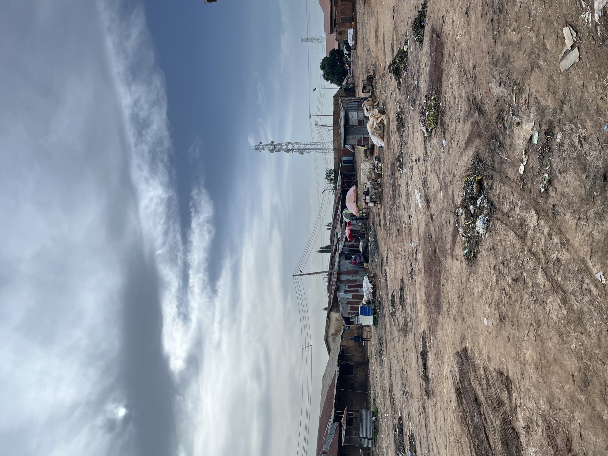 The image depicts an outdoor scene in Gura Top, Nigeria. The foreground is dominated by a dirt ground with scattered debris, suggesting a somewhat unkempt environment. In the mid-ground, there are several structures with corrugated metal roofs, some appearing to be makeshift or informal dwellings and shops. People are visible in and around these structures, some gathered under colorful umbrellas, indicating a marketplace or gathering point. A large telecommunications tower stands prominently in the background, along with other utility poles and power lines. The sky above is partly cloudy with wispy formations. The overall impression is of a busy, lived-in area, possibly a community market or residential zone with limited infrastructure. There is no visible text in the image.