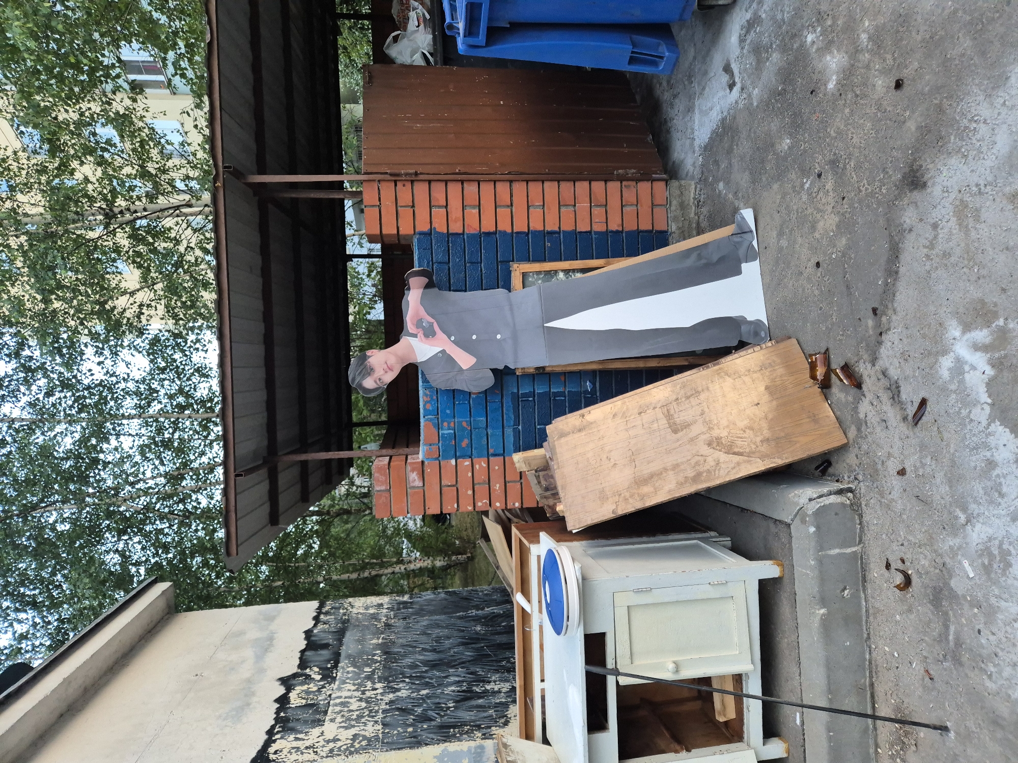 A full-body cardboard cutout of a young East Asian male in a grey suit and white t-shirt stands near a dumpster enclosure. He is posing with his hands forming a heart shape above his chest. The cutout is leaning against a brick wall, half of which is painted blue. To the left of the cutout, there is a white cabinet with a blue circular object on top, and a long black pole resting against it. A large wooden plank is propped up against the cutout and the cabinet. The ground is concrete and appears to be littered with broken glass and debris. In the background, there is a metal roof structure, a brown metal door, a blue dumpster, trees, and a building. The scene appears to be an outdoor area in a city, possibly a residential courtyard.