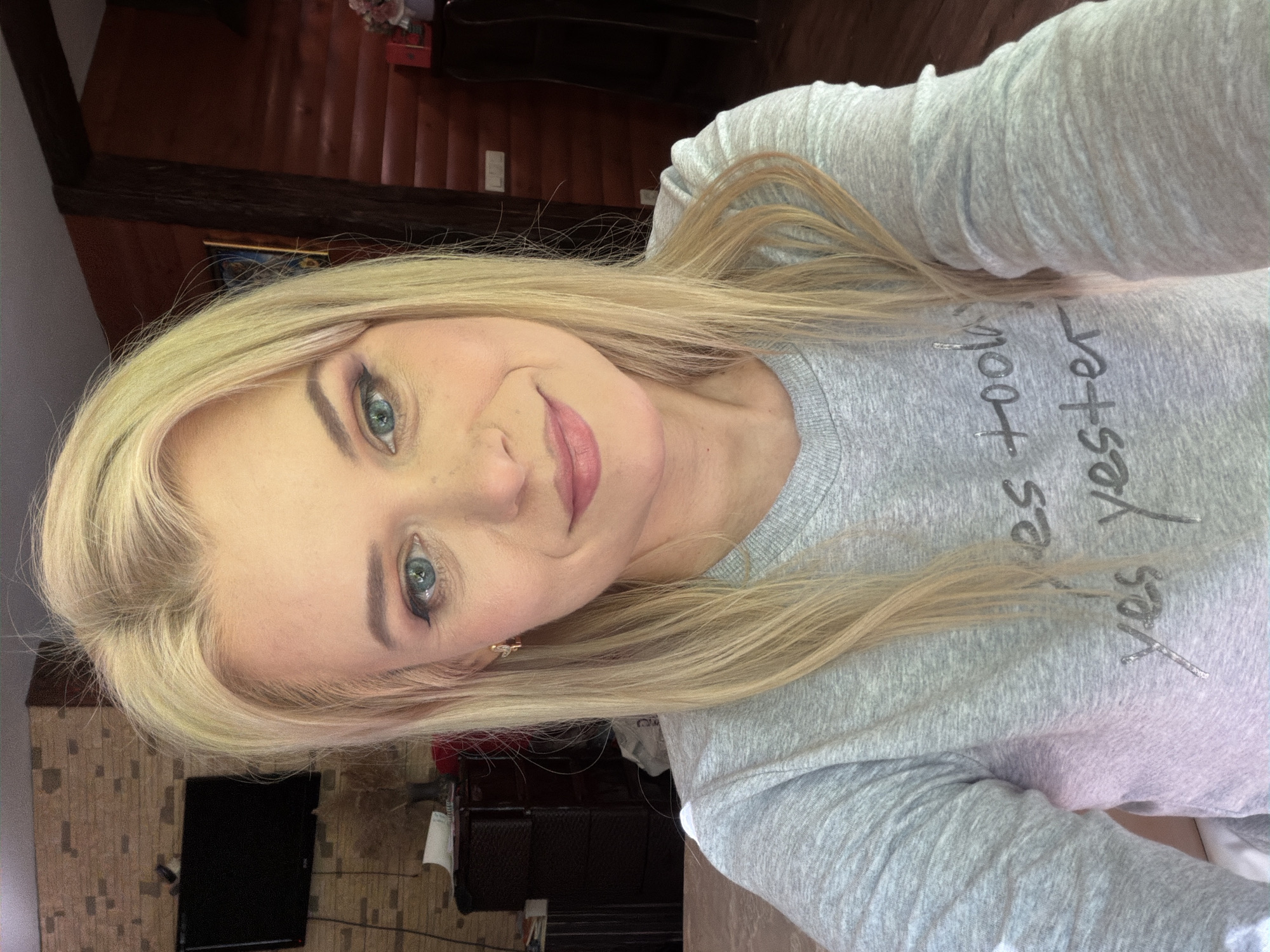 A woman with blonde hair and blue eyes is taking a selfie. She is wearing a gray sweatshirt with the words "yes took yester" printed on it in silver lettering. The background appears to be an indoor setting with wooden beams and a brick wall visible. The lighting suggests it is daytime. The woman's expression is friendly and she is looking directly at the camera. There are some decorative items on shelves in the background, including what looks like a bouquet of flowers.