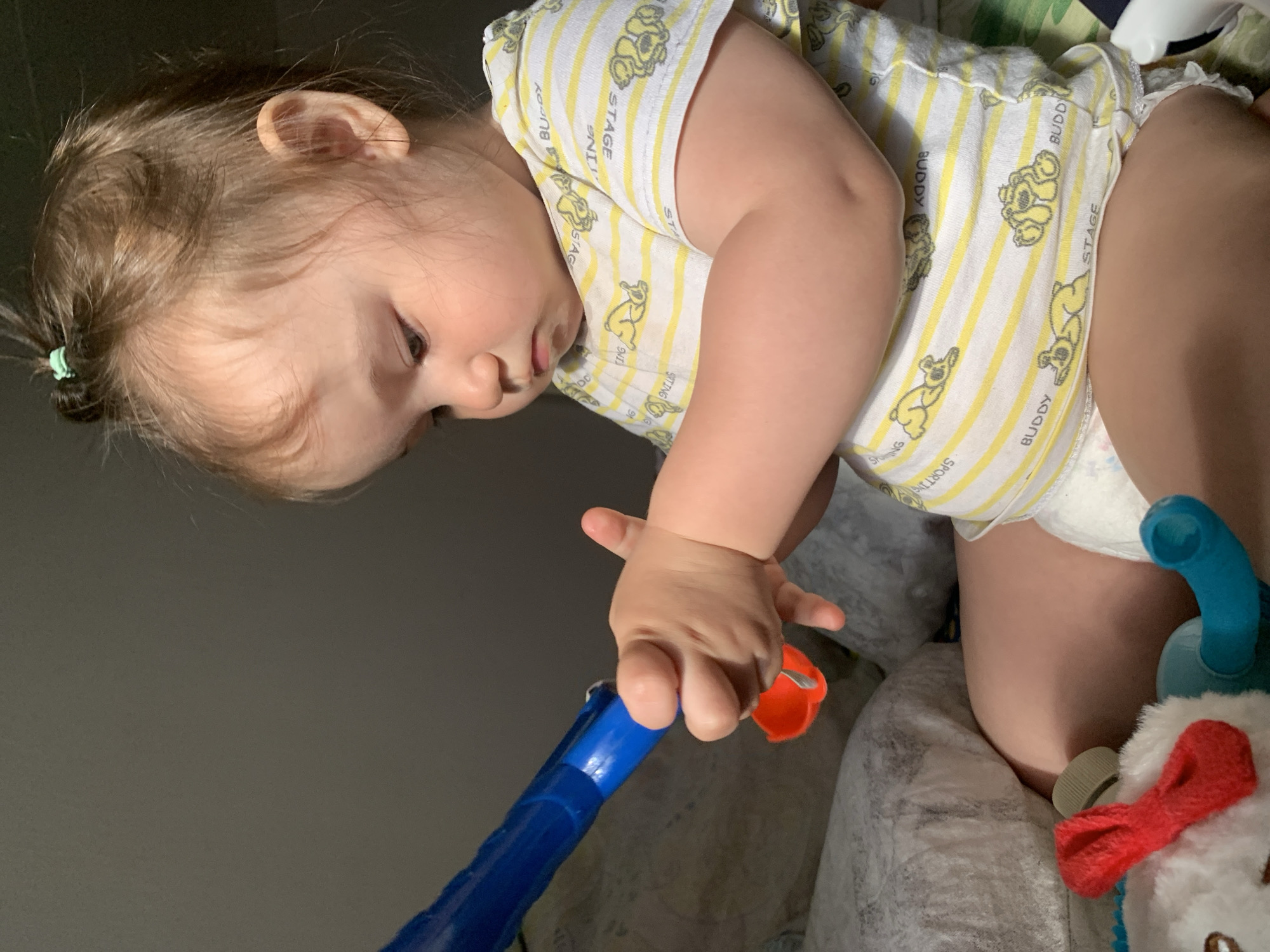 An indoor shot shows a young child sitting and playing with toys. The child, with brown hair pulled into a small ponytail, is wearing a yellow and white striped t-shirt with cartoon bears and the word "BUDDY" printed on it, and a diaper. The child's hands are gripping a blue toy, possibly a wand or a large crayon, with a small orange cup attached. In front of the child, partially visible, is a stuffed animal with a red bow and a blue piece of plastic. The child is positioned on what appears to be a bed or couch with pillows. The lighting suggests a dimly lit room, with light casting shadows on the wall behind the child. The image does not provide any information about the location within the city of Chaikovsky, Russia.