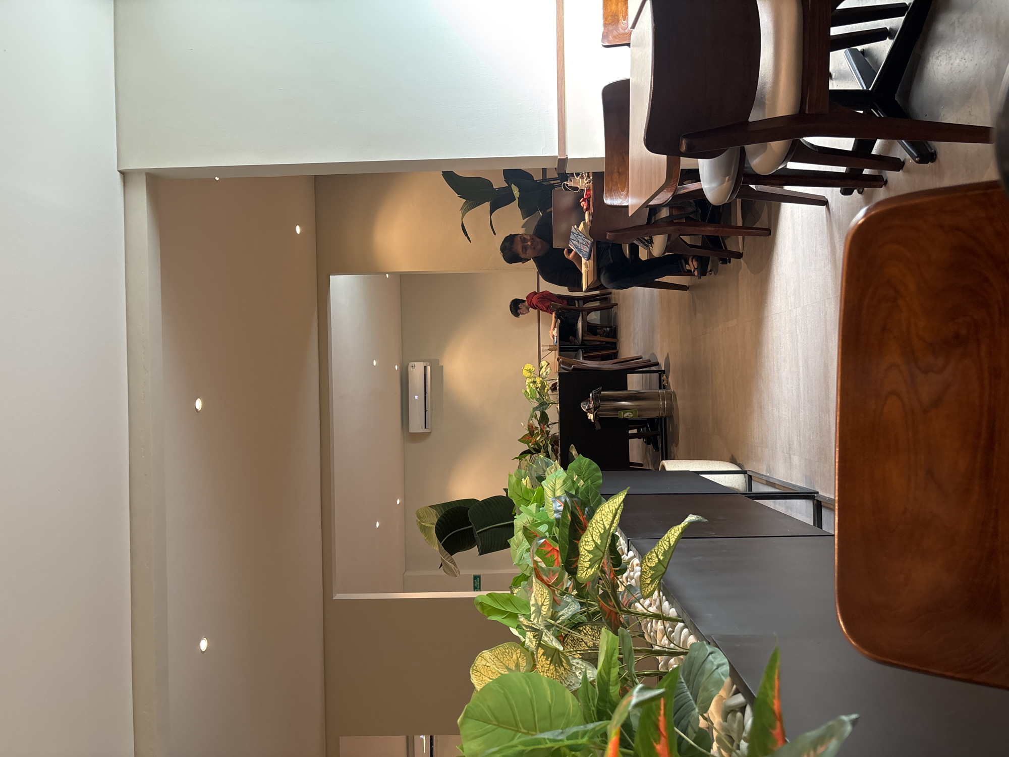 The image depicts the interior of a cafe in West Jakarta, Indonesia. In the foreground, a close-up of a wooden chair is visible. Moving further into the scene, a contemporary cafe interior unfolds. Lush green plants are strategically placed in dark planters, adding a natural element to the space. Several tables with wooden chairs are arranged throughout the cafe.
In the mid-ground, a man is seated at a table, working on a laptop. Another individual, a younger male, is seated further back at a different table. The cafe environment appears calm and conducive to work or relaxation. The lighting is soft, coming from overhead fixtures and natural light filtering in from windows, creating a warm ambiance. The walls are painted in neutral tones, and the floor is tiled. The overall impression is one of a modern and well-maintained cafe.