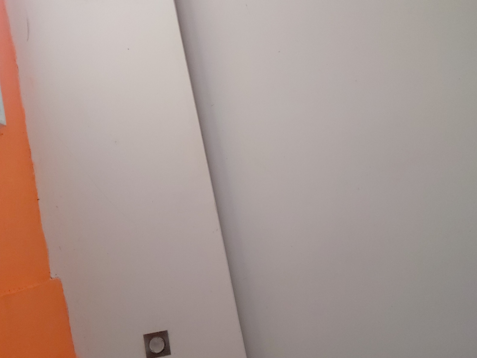 The image displays an indoor architectural detail, featuring a corner or seam where two walls meet. The wall on the left is painted a vibrant, solid orange color, while the larger wall section occupying the right and most of the center is a plain light gray or off-white. A distinct, slightly darker vertical line separates these two color areas, suggesting an interior corner or a panel joint.
Near the bottom left of the image, positioned on the light-colored wall close to the orange section, there is a small, square-shaped fixture with a metallic, circular center. This object appears to be a low-profile light, a switch, or some form of control panel embedded in the wall. The surface of both walls appears smooth.
No people are visible in the scene, and no specific activity is taking place. The lighting is even and diffused, typical of an interior space, making the time of day indistinguishable. There is no visible text. The setting appears to be a modern interior in Santa Ana, Venezuela.