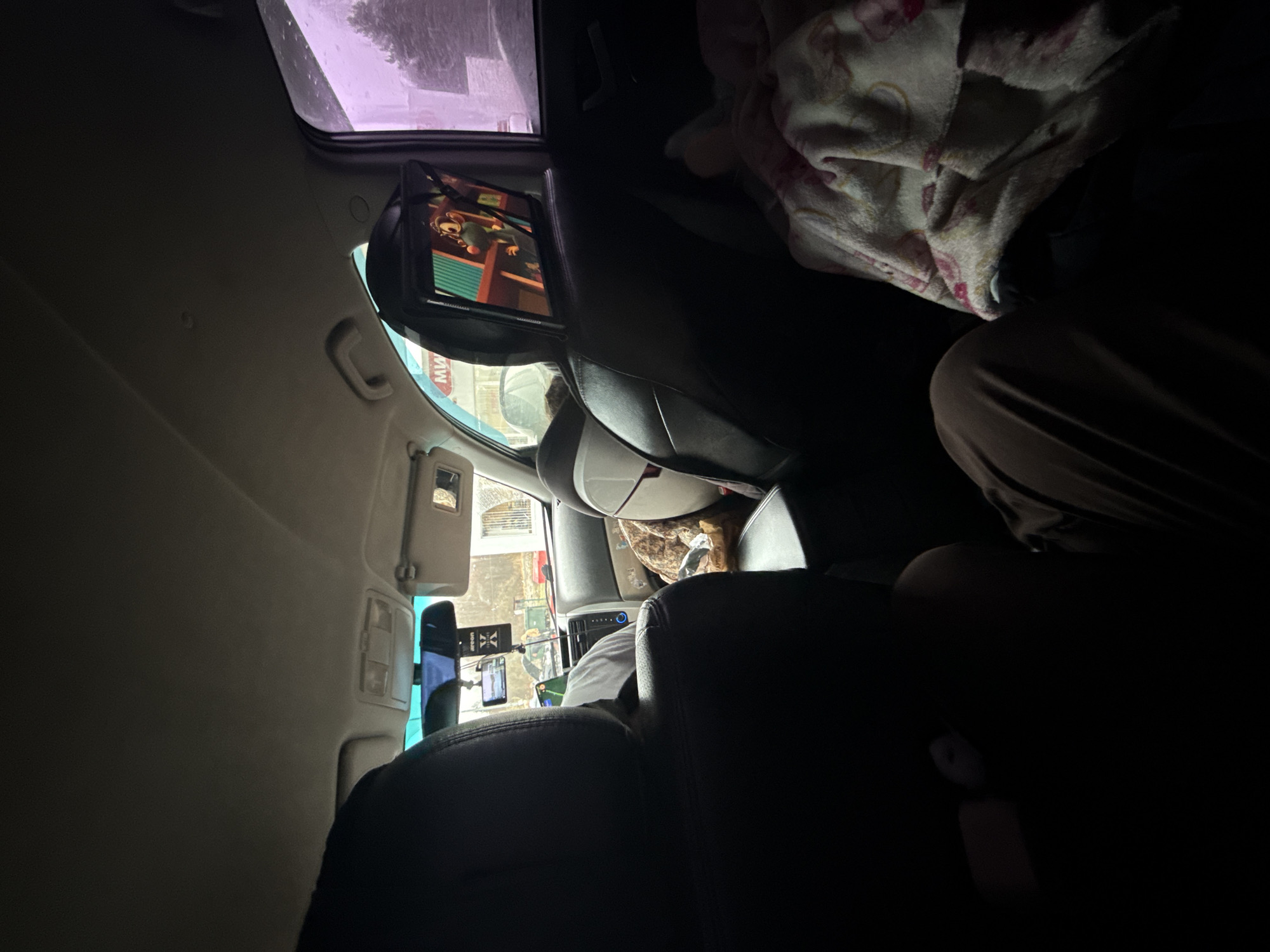 The image captures a unique, counter-clockwise rotated view from the interior of a car, looking forward from a rear passenger seat. Mounted on the back of the front passenger headrest is a tablet displaying an animated character, possibly a squirrel-like creature with large eyes, against a vibrant green and orange background. On the right side of the foreground, a patterned blanket, predominantly white with pink designs, covers what appears to be a seat or items, with a person's legs wearing brown pants visible beneath it.
Further ahead, the back and shoulder of a person in the front passenger seat, wearing a light-colored shirt, are partially visible. The car's dashboard, central console, and a rearview mirror are discernible. Attached near the rearview mirror is a black rectangular object, possibly a car air freshener. Several phone mounts or electronic devices, one displaying a map or navigation screen, are fixed around the dashboard area.
Outside the car, daylight streams through the windows, revealing urban or suburban buildings and hints of foliage. A faint red "MW" or "HW" text is visible on a sign outside, through the lower-left side window. The scene depicts a daytime journey or stop in a vehicle, set in Kiselevka, Russia.