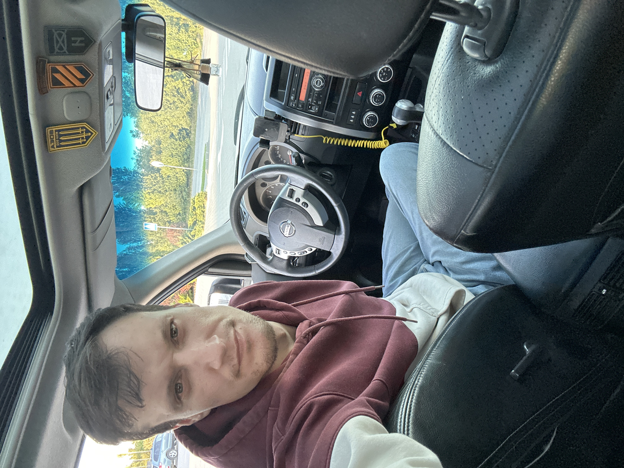 A man with short brown hair and a light beard is sitting in the driver's seat of a car. He is wearing a maroon and white hoodie and light blue jeans. He is looking directly at the camera. The car interior is visible, including the dashboard, steering wheel, and gear shift. There are three patches on the car's ceiling, one yellow with three swords, one orange with black stripes, and one green with a black symbol. A rearview mirror hangs from the ceiling, with a small decorative item attached. The exterior of the car is partially visible through the windshield, showing a parking lot and trees. The scene is set in the city of Kamianets-Podilskyi, Ukraine.