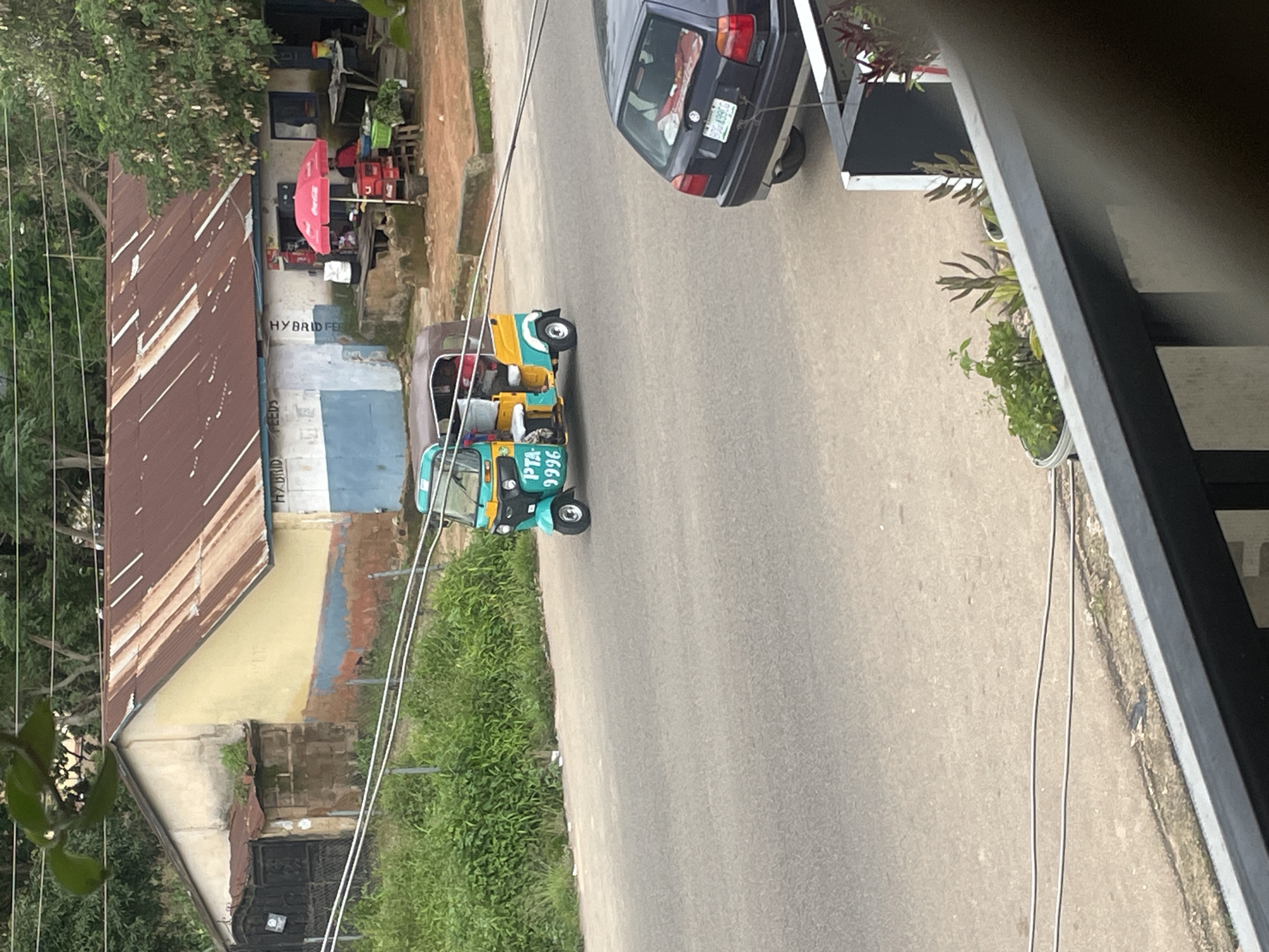This is an eye-level, medium shot capturing a street scene in Jos, Nigeria. A teal and yellow auto-rickshaw, also known as a "Keke Napep," is the central focus, traveling on a paved road. The auto-rickshaw has "PTA 9996" written on its side. To the right of the auto-rickshaw, a dark-colored hatchback car is parked, facing away from the viewer.
In the background, a building with a rusted corrugated iron roof and a pale yellow facade is visible. The building appears to be a shop or a market stall, with a red Coca-Cola umbrella set up outside. There are people visible under the umbrella, and goods are displayed on tables. A red cooler box is also present. A lush green fence runs along the side of the road. The environment suggests a semi-urban setting, typical of parts of Jos.