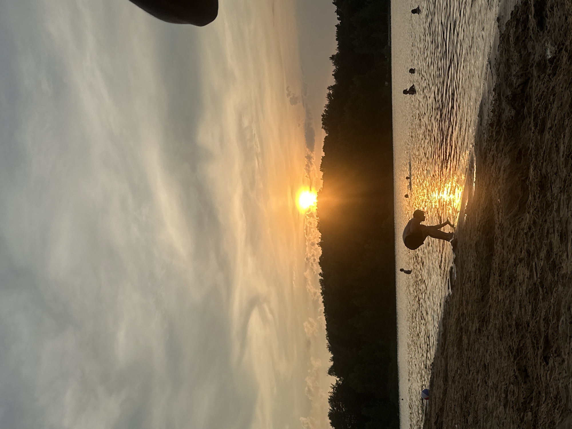 An image depicts a sunset over a body of water, with a sandy beach in the foreground and a treeline in the background. Several people are visible in the water, some appearing to be swimming, while others are standing. One person is on the beach, bent over near the water's edge. A beach ball is also visible on the sand. The sun is low on the horizon, casting a warm glow across the scene and reflecting on the water. The sky is partly cloudy with a mix of gray and orange hues. The location is identified as Lytkarino, Russia.
