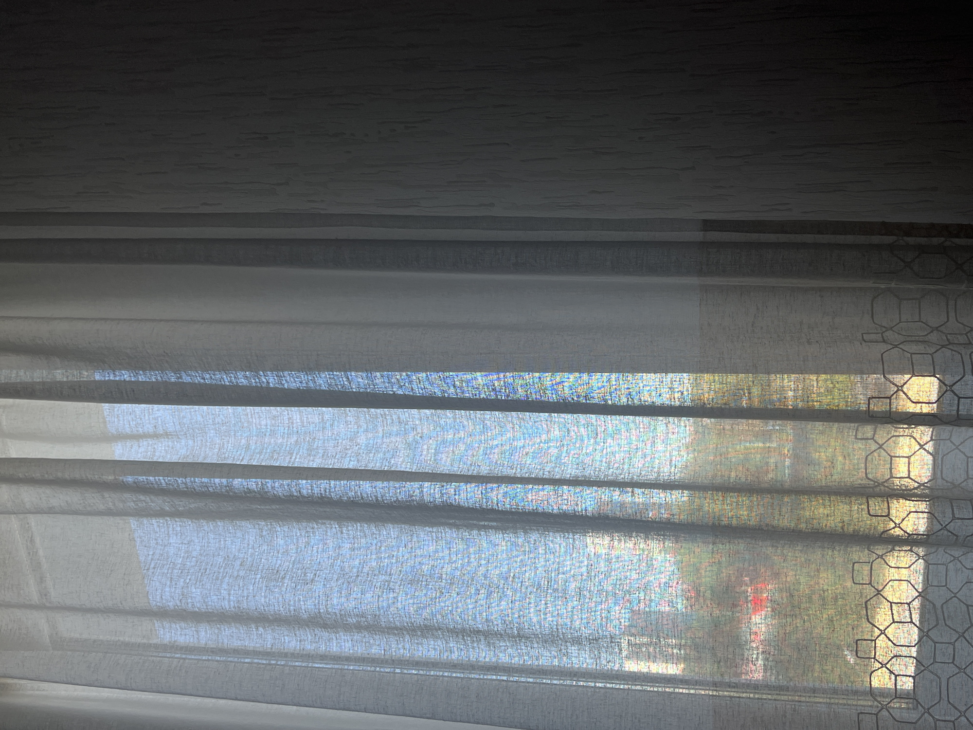 The image shows a view through a window covered with sheer, light-colored curtains. Outside, the blurred impression suggests a cityscape with greenery and a possible red object (likely a car or building detail). The specific location within Perm, Russia, cannot be determined from this image. The focus is on the interior view obstructed by the curtain, leaving the external details indistinct.