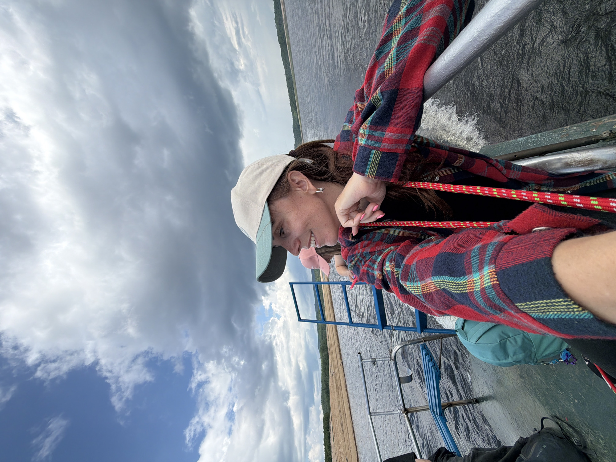 A woman wearing a plaid shirt and a baseball cap is on a boat. She is looking to her right and smiling. She has long brown hair. In the background, there is a body of water and land with trees. The sky is cloudy.
