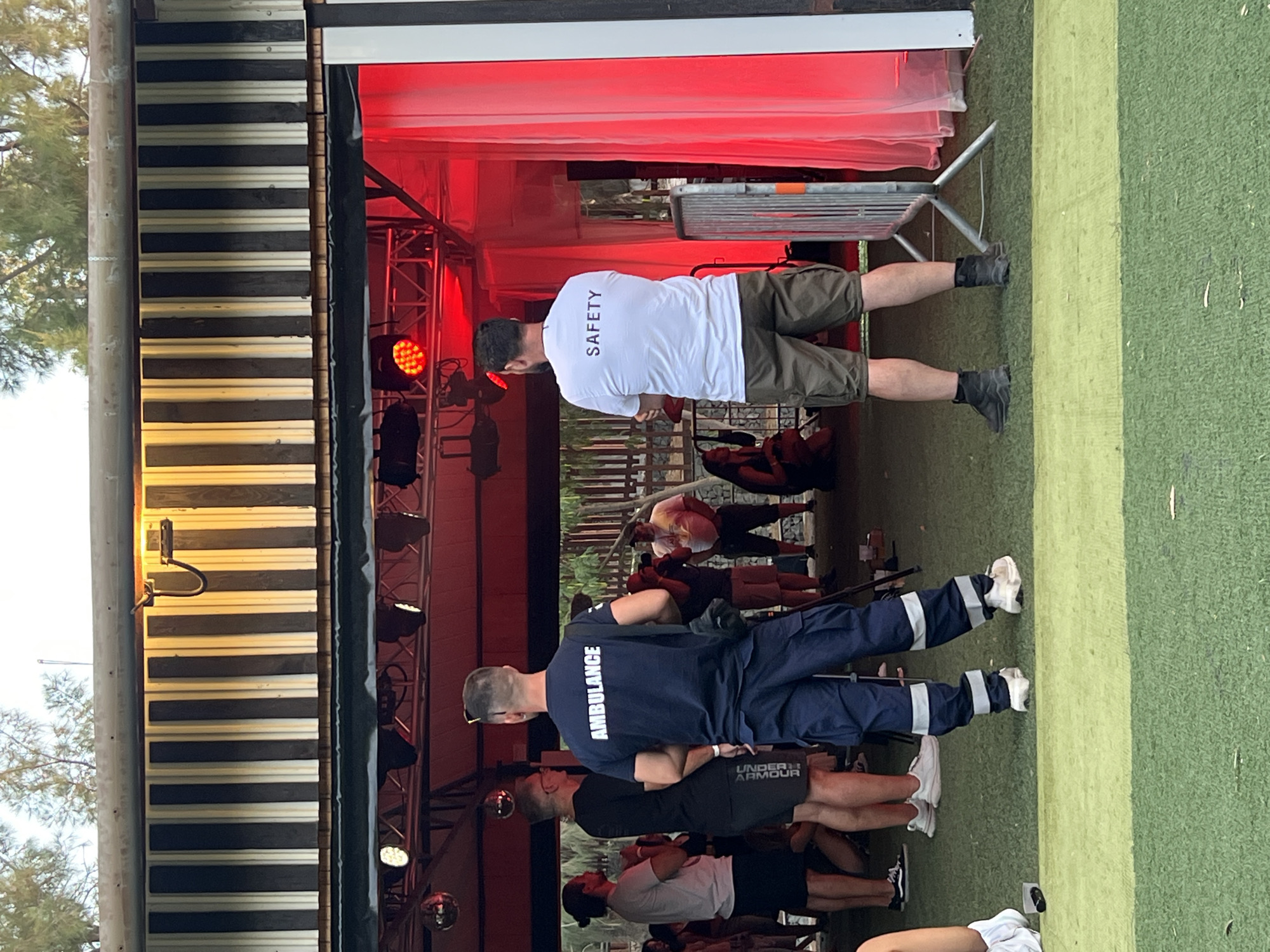 The image depicts an outdoor event, possibly a festival or concert, in Analiontas, Cyprus. Two individuals in uniforms stand facing away from the camera. The person on the left wears a dark blue shirt with "AMBULANCE" printed in white on the back, and matching dark blue cargo pants with reflective stripes on the lower legs. The person on the right wears a white t-shirt with "SAFETY" printed in black on the back, and khaki cargo shorts. They are standing on a green artificial turf surface in front of an open structure with a black and white striped facade above. The structure's interior is lit with red lighting. In the background, other people are visible, some standing and some seated, along with trees and fencing. A metal barrier is positioned to the right of the "SAFETY" uniformed individual.