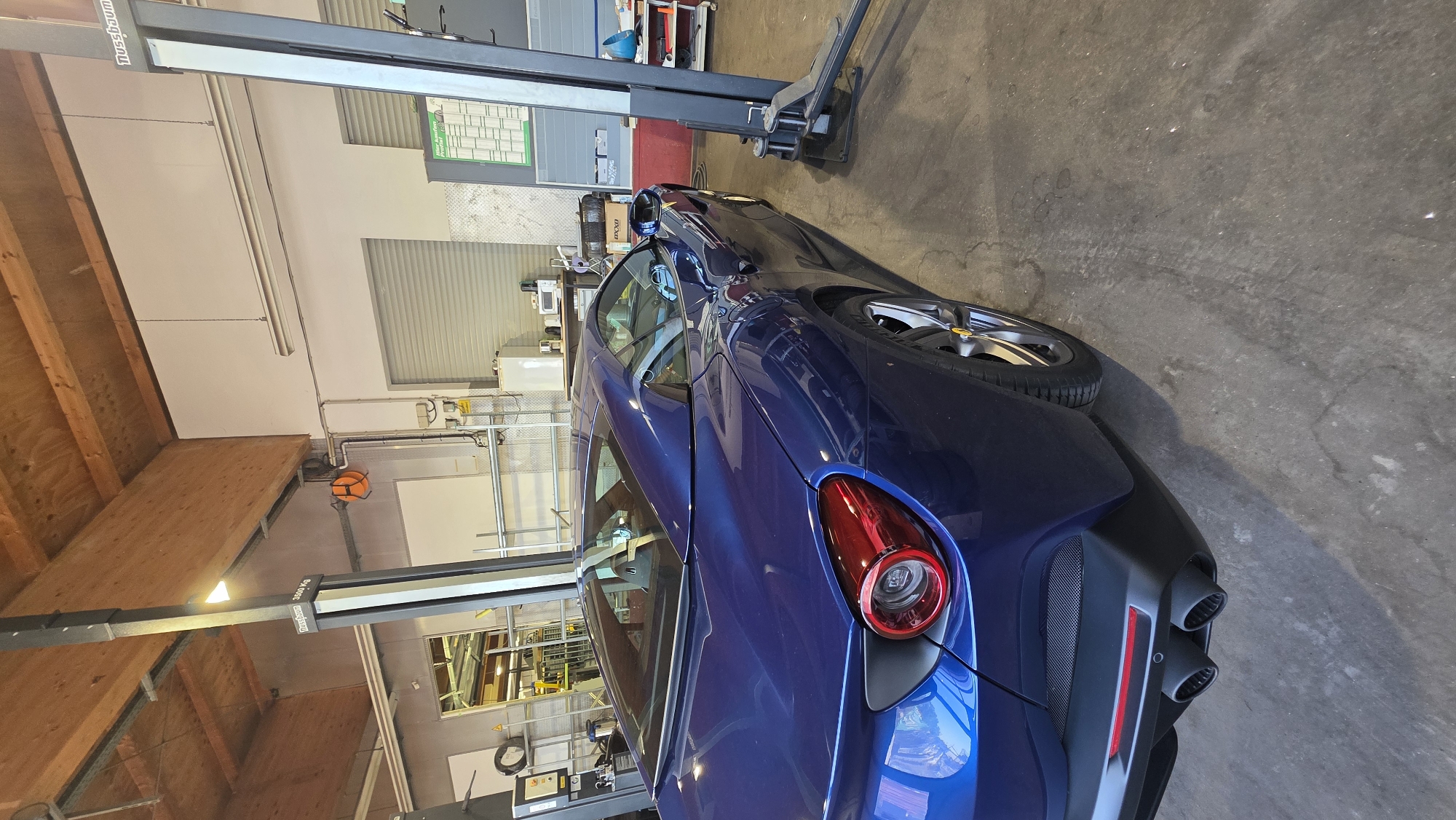 A blue Ferrari is parked in a garage with a hydraulic lift next to it. The car is facing away from the camera, and its rear end is visible. The garage appears to be a workshop, with tools and equipment visible in the background. The lighting is dim, and the overall impression is one of a utilitarian space.