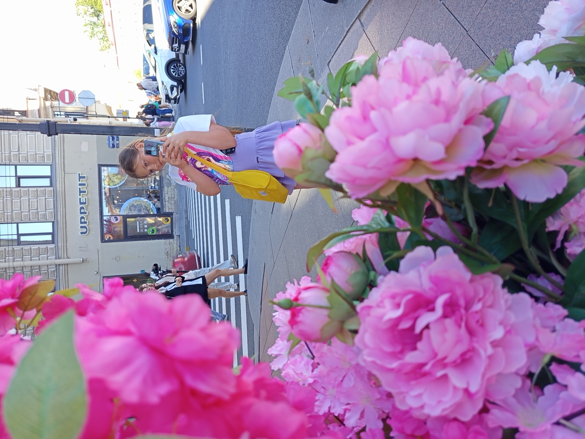 A young girl stands on a sidewalk in Saint Petersburg, Russia, holding a smartphone and taking a selfie. She is wearing a white t-shirt with a colorful graphic and a light purple skirt, and carrying a yellow bag over her shoulder. In the background, people are crossing a pedestrian street, and cars are parked on the side of the road. A building with the sign "UPPETIT" is visible behind the girl, and a street sign indicating "GOROKHOVAYA" can be seen. The foreground is blurred with large pink flowers, likely peonies, obscuring the lower part of the image. The scene appears to be a bright, sunny day.