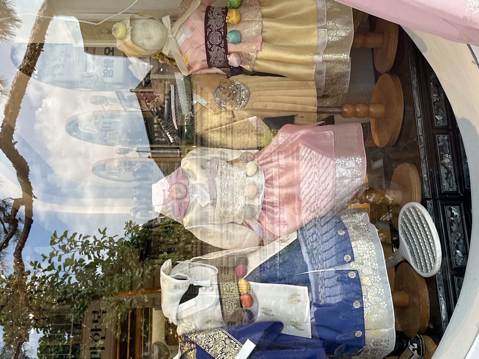 This image displays a store window in Jeonju-si, South Korea, exhibiting traditional Korean clothing known as Hanbok. The mannequins are adorned with colorful and intricately designed Hanbok, featuring bright blues, pinks, and yellows, with gold and silver embroidery. The clothing includes the characteristic jeogori (upper garment) and chima (skirt) or baji (trousers), along with decorative sashes and accessories such as colorful decorative knots and pearl necklaces. Reflections in the window suggest an outdoor environment with trees and a sky, possibly indicating the presence of traditional Korean architecture in the background. The window also shows a glimpse of the store's interior, with wooden display stands and a dark, ornate base for the mannequins. A white, oval-shaped object with a grid pattern, resembling a badminton racket or a sieve, rests on the floor in front of the mannequins. The scene is brightly lit, highlighting the details of the Hanbok.