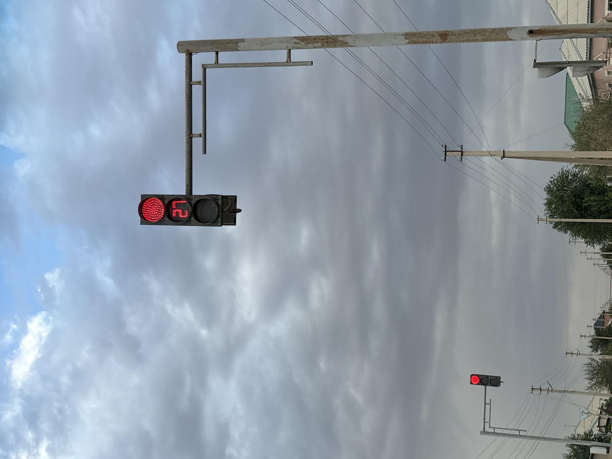 A red traffic light with a countdown timer showing "27" is illuminated. The traffic light is attached to a metal pole which is part of a larger traffic signal assembly, positioned over a road. In the distance, another traffic light, also showing red, is visible. Power lines and poles are present throughout the scene. The sky is overcast with heavy clouds. The setting appears to be a street in Рахат ауылдық әкімдігі, Kazakhstan, with houses and trees visible on either side of the road.