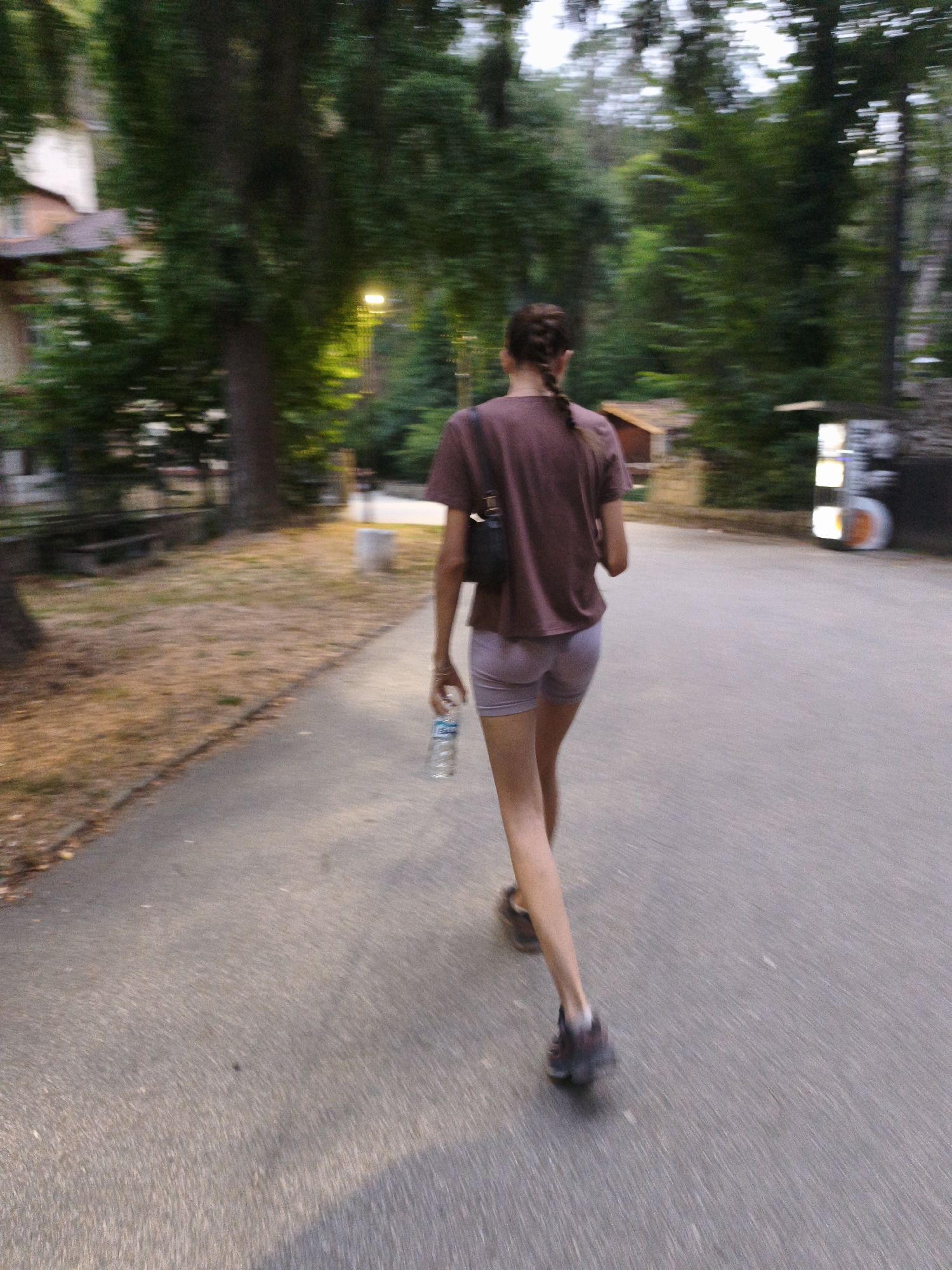 A person with braided brown hair wearing a maroon t-shirt and light purple shorts walks away from the camera on a paved path. They are holding a clear plastic bottle of water in their right hand. The path is bordered by a grassy area with fallen leaves on the left and trees on the right. In the background, a building with a light-colored roof is visible amongst the trees. The scene appears to be a park or residential area in Lovech, Bulgaria, given the presence of a concrete bollard and a sign with illuminated panels in the background. The image has motion blur, indicating movement.