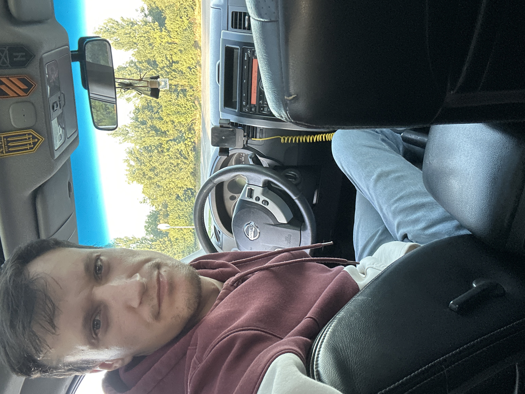 A man is sitting in the driver's seat of a Nissan car, looking at the camera. The car's interior is visible, including the dashboard, steering wheel, and seats. A phone mount with a phone is attached to the dashboard. The car appears to be parked on a road, with trees and foliage visible through the windshield. The scene is set in Kamianets-Podilskyi, Ukraine.