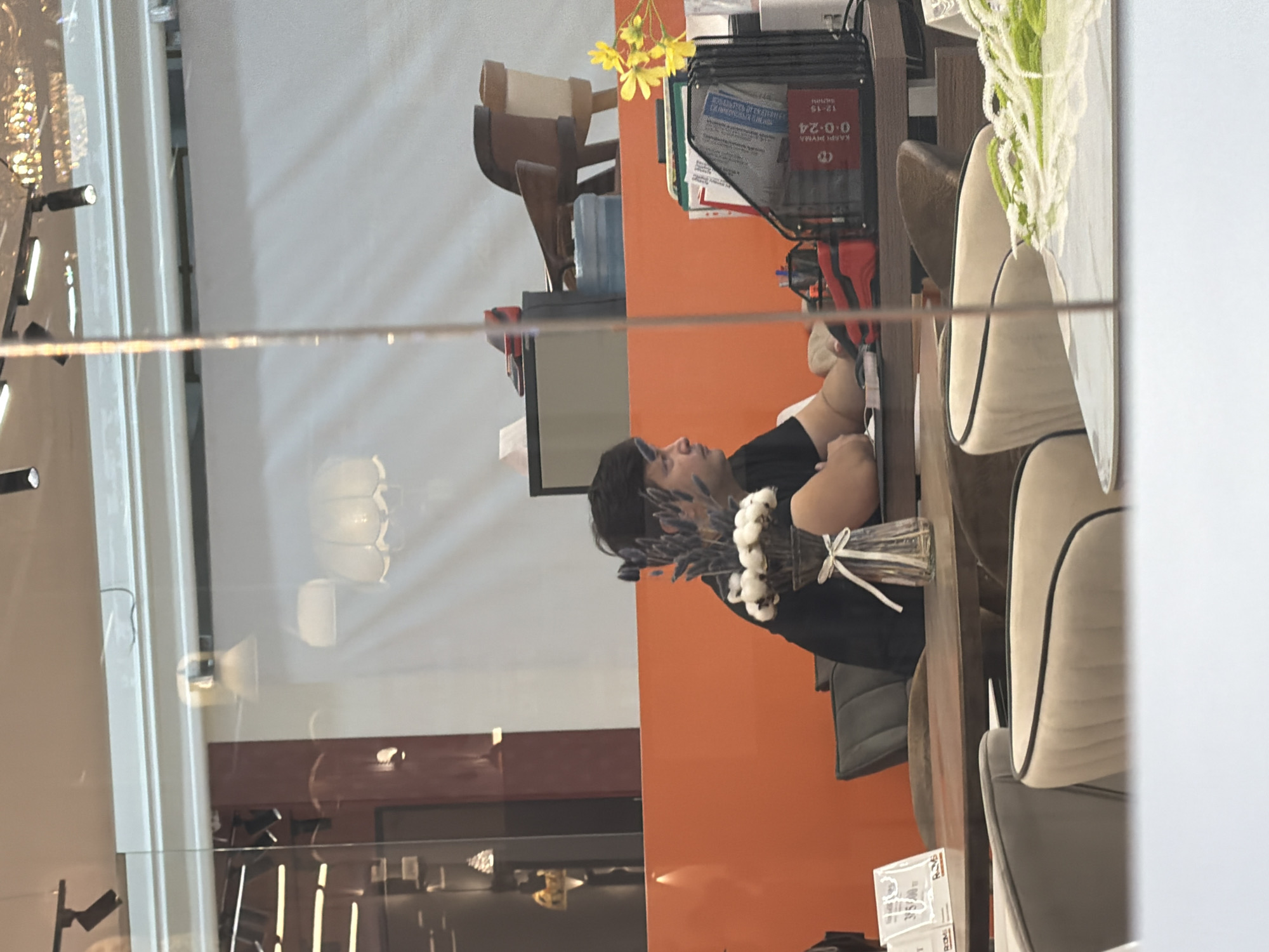A man in a black t-shirt is seated at a wooden table, facing to the right. He has dark hair pulled back in a bun and is wearing blue eye makeup. In front of him is a vase with white cotton-like flowers and a decorative ribbon tied around it. To his right, on the table, is a computer monitor and a black wire mesh organizer filled with papers. A red stapler and a blue water bottle are also visible on the table. The background features an orange wall and a wooden chair. The scene appears to be indoors, possibly in a cafe or office space, in Astana, Kazakhstan, as indicated by the presence of Cyrillic text on some of the documents and the overall aesthetic.