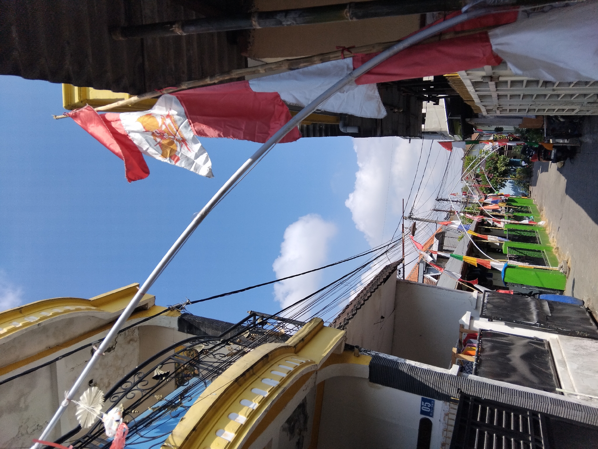 In Kepuh Kiriman, Indonesia, a street scene is depicted on a bright, sunny day with a clear blue sky and scattered white clouds. The foreground is dominated by a large, yellow building with a curved balcony railing. To the right, a bamboo structure and corrugated metal roof suggest a building or awning.
Several red and white Indonesian flags are prominently displayed. One large flag, featuring the Garuda Pancasila emblem, is mounted on a pole attached to the bamboo structure. Smaller triangular flags in red, white, green, and yellow are strung along the street, adorning fences and poles.
The street itself is paved and appears to be lined with residential buildings. Black metal fences and green painted walls are visible along the sides of the road. In the distance, more houses and trees can be seen. A parked motorcycle is present on the right side of the street. The presence of numerous flags suggests a celebration or national holiday. The overall impression is one of a vibrant and patriotic community setting.