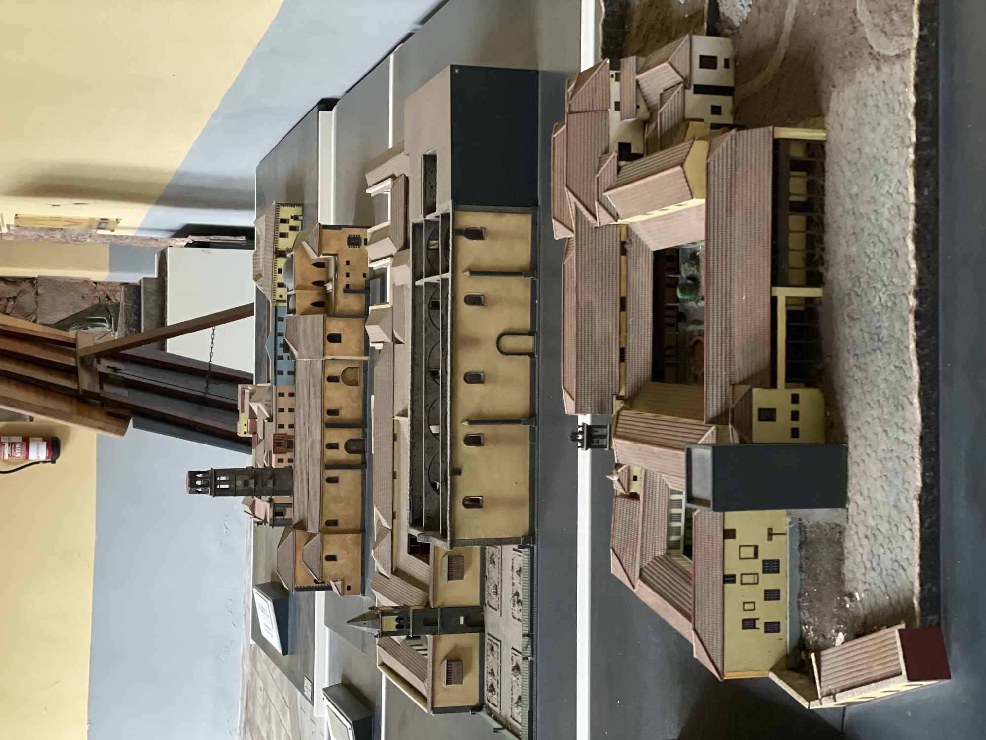 This media file presents an intricately detailed architectural model of a city, likely San Cristóbal De La Laguna, Spain, viewed from an elevated, slightly oblique perspective. The model features numerous buildings constructed from materials resembling light beige or yellow walls with reddish-brown tiled roofs, typical of historical European architecture. Many structures also incorporate dark flat roofs, suggesting different architectural styles or modern additions within the representation.
A prominent feature is a tall, dark tower with several levels and arched openings, standing amidst the densely packed miniature buildings. Streets are depicted in a dark grey color, with lighter grey elements possibly representing sidewalks or road markings. Other areas within the model show textured ground, some appearing cobbled or unpaved. The scale of the model suggests a significant level of detail, showcasing a complex urban layout with various building types and configurations, including courtyards and open spaces.
In the background, outside the model's immediate area, an indoor setting is visible. This includes a yellow painted wall, a grey painted wall, and some wooden structures or beams. A red fire extinguisher with unreadable white text is affixed to the yellow wall. No specific activity or event is taking place within the static model; it serves as a visual representation of the city's architecture and layout. The lighting appears to be artificial, indicative of an exhibition or display setting.