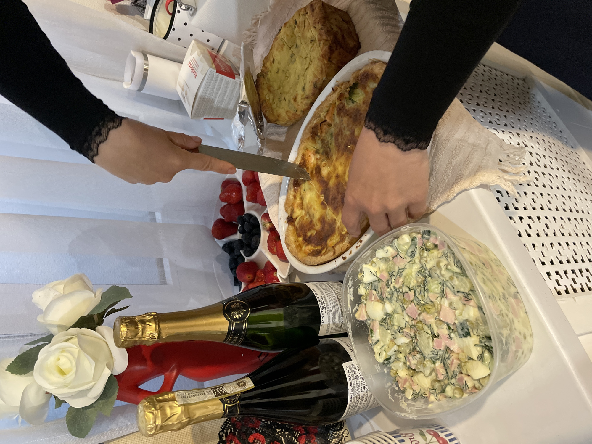 Here is a description of the image:
The image shows a celebration with food and drink. Two individuals with black sleeves are seen preparing food on a white surface. One is cutting a baked dish, potentially a quiche, with a knife. Nearby is a loaf of bread-like food. There are strawberries and blueberries. Two bottles of sparkling wine lie in the foreground among some white roses on a red object. Next to the champagne is a large container of what appears to be Russian salad ("Olivier").
Labels on the bottles indicate that the beverages are sparkling wines, specifically "MOSSIERE ASTI" and "VEUVE ALBAN BRUT".
There's a box labeled "Konfesta" visible.
The location "Sochi, Russia" cannot be confirmed from the details in the image.