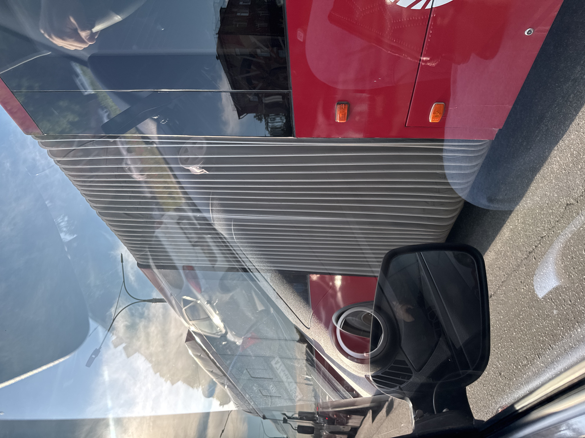 A bus is visible in the image, specifically its accordion-like articulated section. The bus has a red panel with orange lights, and its reflective window shows building reflections. The image seems to be taken from inside another vehicle, evident from the car's side mirror and interior components. The location is Perm, Russia, based on the license plate visible on the vehicle.