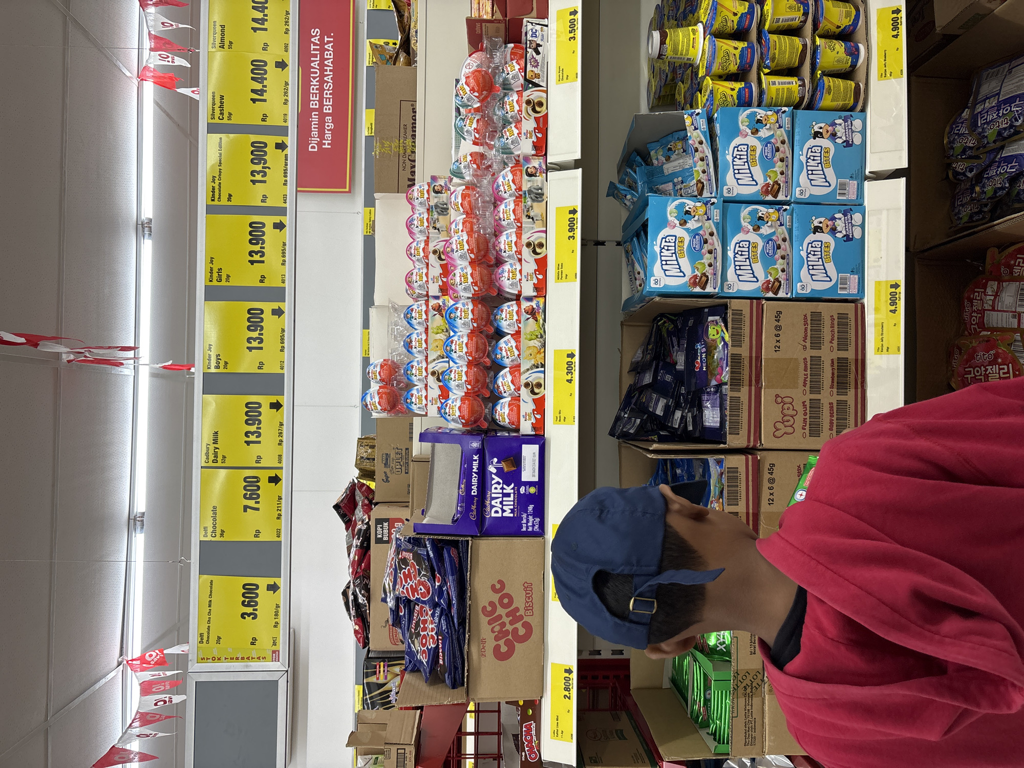 A person wearing a red hooded sweatshirt and a blue baseball cap is standing in a supermarket aisle. The aisle is stocked with various confectionery products such as Kinder Joy eggs, Milikta Bites, Yupi sweets, Delfi chocolates, and Cadbury Dairy Milk chocolate. Price tags with the local currency (Rp) are visible next to the products, indicating the cost of each item. The supermarket aisle is well-lit, and red and white bunting is hung along the top.