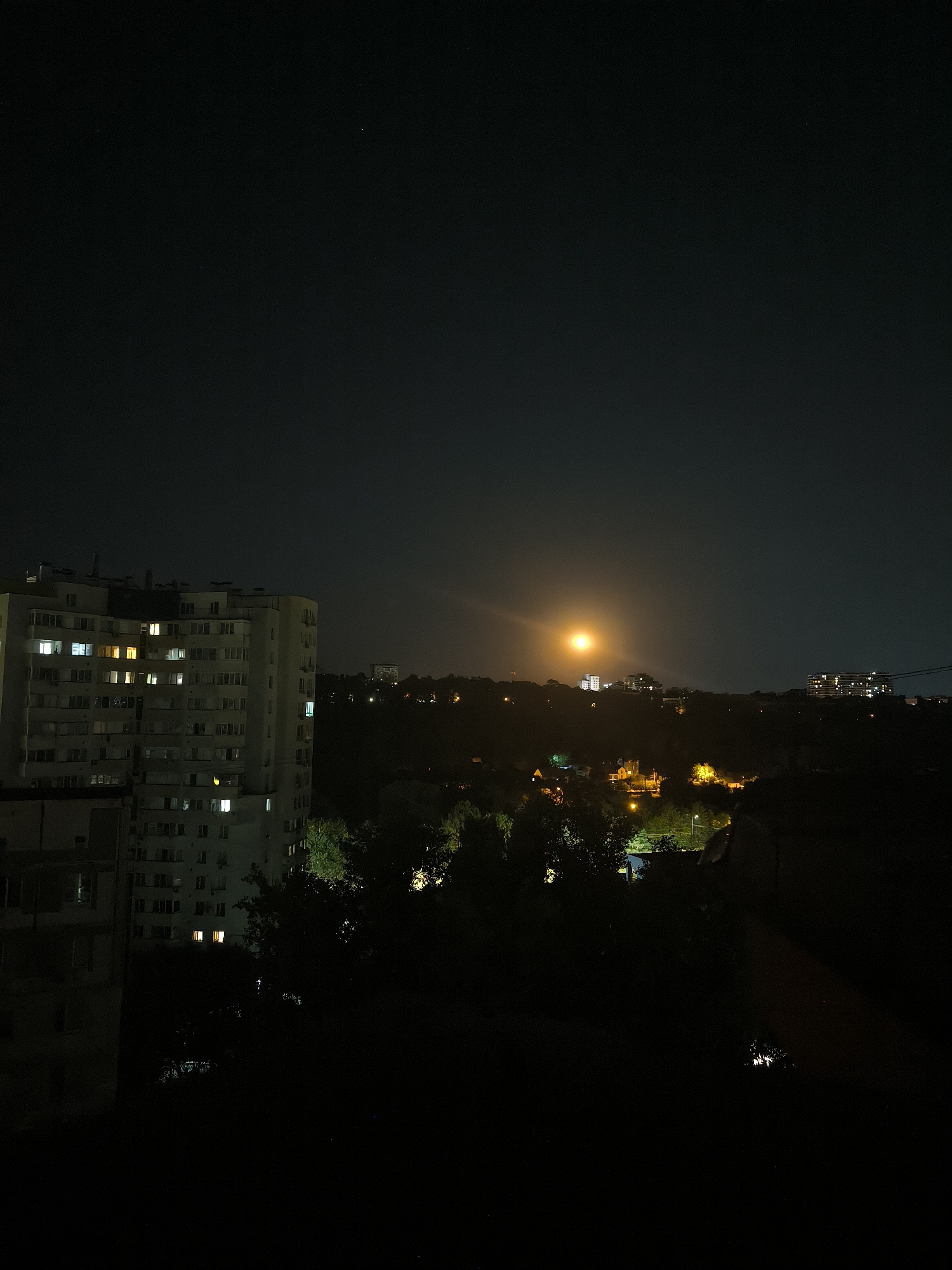 The image depicts a nighttime cityscape in Chișinău, Moldova. In the foreground, on the left, is a large, multi-story apartment building with many windows illuminated by interior lights. To the right of this building, a dark, hilly landscape is visible, dotted with trees and houses, also showing some illuminated windows. In the middle distance, a cluster of buildings stands on a hill, with a bright, orange-yellow orb, possibly the moon or a strong light source, positioned in the sky above them, creating a significant glow. Further in the distance, more buildings and lights can be seen, suggesting a more densely populated area. The sky is dark, with a few faint stars visible. The overall scene conveys a sense of urban life at night, with the artificial lights of the city contrasting with the darkness of the sky.