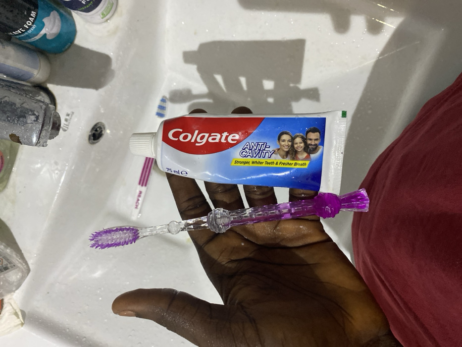 The image captures a close-up, eye-level view of a person's dark-skinned hand holding a tube of Colgate toothpaste and a purple toothbrush over a white bathroom sink. The hand appears wet, suggesting recent use of water or preparation for hygiene.
The main subjects are the hand, the Colgate toothpaste, and the purple toothbrush. The toothpaste tube is white and blue, featuring the "Colgate" logo prominently in red. Below the logo, it reads "ANTI-CAVITY" with the tagline "Stronger, Whiter Teeth & Fresher Breath." A small image of a family (a woman, a child, and a man) is visible on the tube. The volume "75 mle" is also printed on it. The purple toothbrush, held horizontally beneath the toothpaste tube, has clear plastic bristles and a patterned handle.
In the background, within the sink basin, several other toiletries are visible. To the left, a silver faucet shows signs of wear, and next to it are bottles, including one partially visible with "AVE FOAM" printed on it, likely shaving foam. Another toothbrush, pink and white with "REACH" visible on its handle, lies in the sink to the left of the hand. The sink itself is white and appears wet, with some water droplets and possibly soap residue. A dark drain hole is visible in the center of the sink. Strong overhead lighting casts distinct shadows of the bottles and other objects onto the sink's surface, creating a somewhat stark contrast. On the far right of the frame, a portion of a red garment is visible, likely clothing worn by the person holding the items.
The scene depicts a common morning or evening routine, likely in a residential bathroom in Lakka, Sierra Leone, as indicated by the context. The items are arranged as if about to be used for dental hygiene.