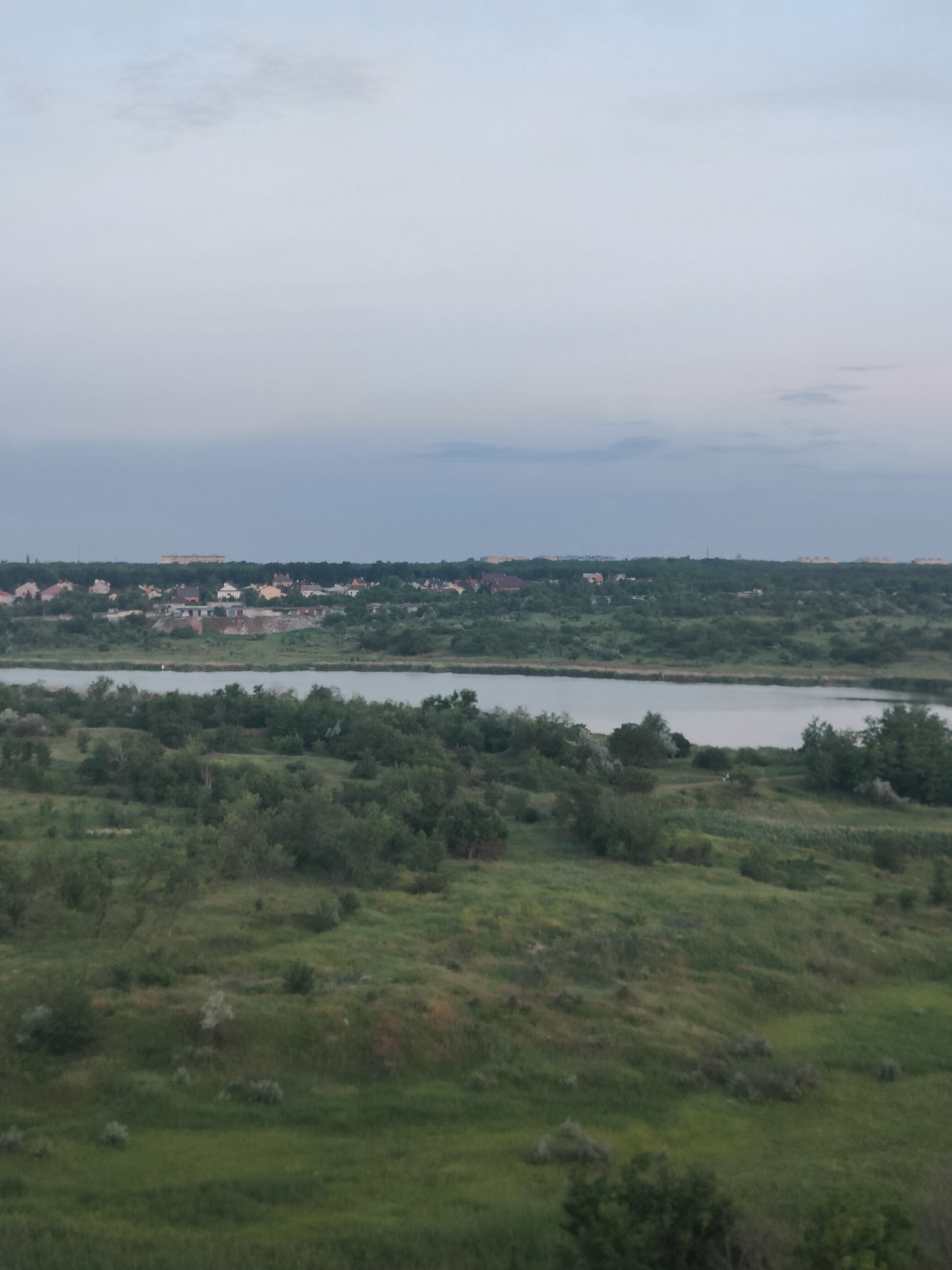 An aerial, long-shot photograph captures a serene, pastoral landscape under a pale blue, overcast sky. The scene is dominated by lush green vegetation, with rolling hills covered in grass and scattered trees. A tranquil body of water, possibly a river or lake, meanders through the middle ground, reflecting the muted tones of the sky. In the distance, a small cluster of houses with red and brown roofs dots the horizon, nestled amongst the dense greenery. The architecture of these houses suggests a rural or suburban setting. The overall impression is one of quietude and natural beauty. The location is the city of Kryvyi Rih, Ukraine.