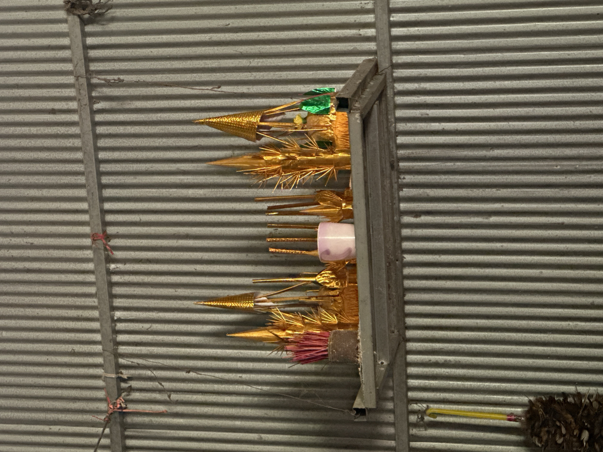 The image shows a shelf mounted on a corrugated metal wall in Thbong, Cambodia. On the shelf are various offerings and decorations, predominantly gold and pink. A bundle of pink incense sticks sits in a woven container on the left. To the right of the incense are several cone-shaped gold ornaments of varying sizes, some with intricate detailing. A small pink cup is also present, and further to the right are more gold decorations and two bright green decorative objects. The shelf itself appears to be made of unfinished wood or metal. A yellow object with a feather-like decoration hangs on the wall to the left of the shelf. The overall scene suggests a religious or memorial display.