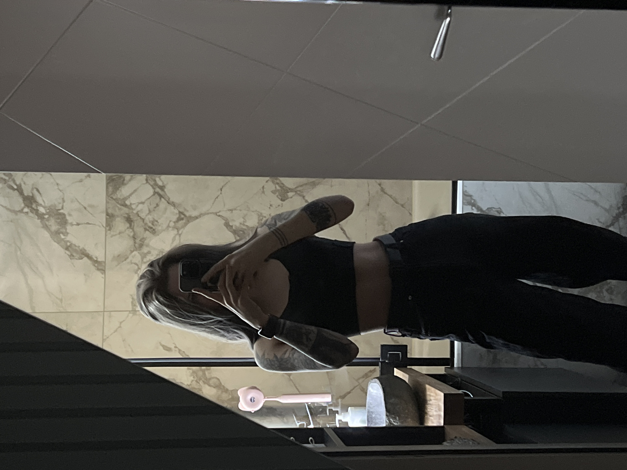 A woman is taking a mirror selfie in a bathroom. The woman has gray hair and tattoos on her arms. She is wearing a black crop top and black pants. The bathroom has gray ceiling tiles and marble-patterned wall tiles. There is a shelf with various items on it, including a pink object shaped like a bear. The specific location in the city of Khimki, Russia is impossible to determine from this image.