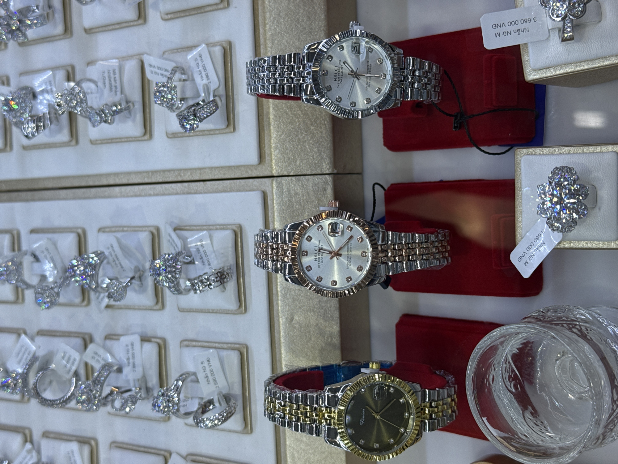 The image captures a display case in a jewelry store, likely located in Tay Nha Trang Ward, Vietnam, judging by the Vietnamese currency (VND) listed on the price tags. The case is filled with an assortment of jewelry, primarily diamond rings and wristwatches.
On the left side of the frame, several trays showcase diamond rings of various designs. Each ring is mounted on a small display card with a price tag attached.
On the right side, a selection of wristwatches is presented on red velvet displays. Three watches are prominently visible: one with a silver-colored band and face, another with a two-tone (silver and gold) band and a silver face, and a third with a two-tone band and a dark green face. Each watch appears to be a dress or formal style. The text on one watch face shows “HALEI”. The dial face of the dark green watch appears to display the text “Dinimi”. The display also features a glass that is engraved.
The lighting is bright and the focus is sharp, allowing for clear visibility of the jewelry and price tags. The overall impression is that of a high-end retail environment.