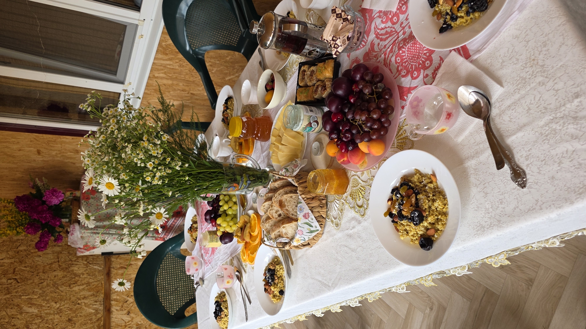 A table covered with a white tablecloth is set for a meal in what appears to be an outdoor or semi-outdoor setting, possibly a veranda or gazebo. The background is a wooden wall. On the table, there are several plates of food, including what looks like oatmeal or porridge topped with dried fruits and nuts. There are also bowls of fruit, including grapes and apricots, sliced oranges, a basket of bread, jars of jam, a container of coconut oil, and a French press coffee maker. A bouquet of daisies is prominently displayed in a vase in the center of the table. The table is also adorned with a decorative runner. Two green plastic chairs are visible on either side of the table. The lighting suggests it is daytime. There are no people visible in the image. The scene depicts a leisurely breakfast or brunch.
