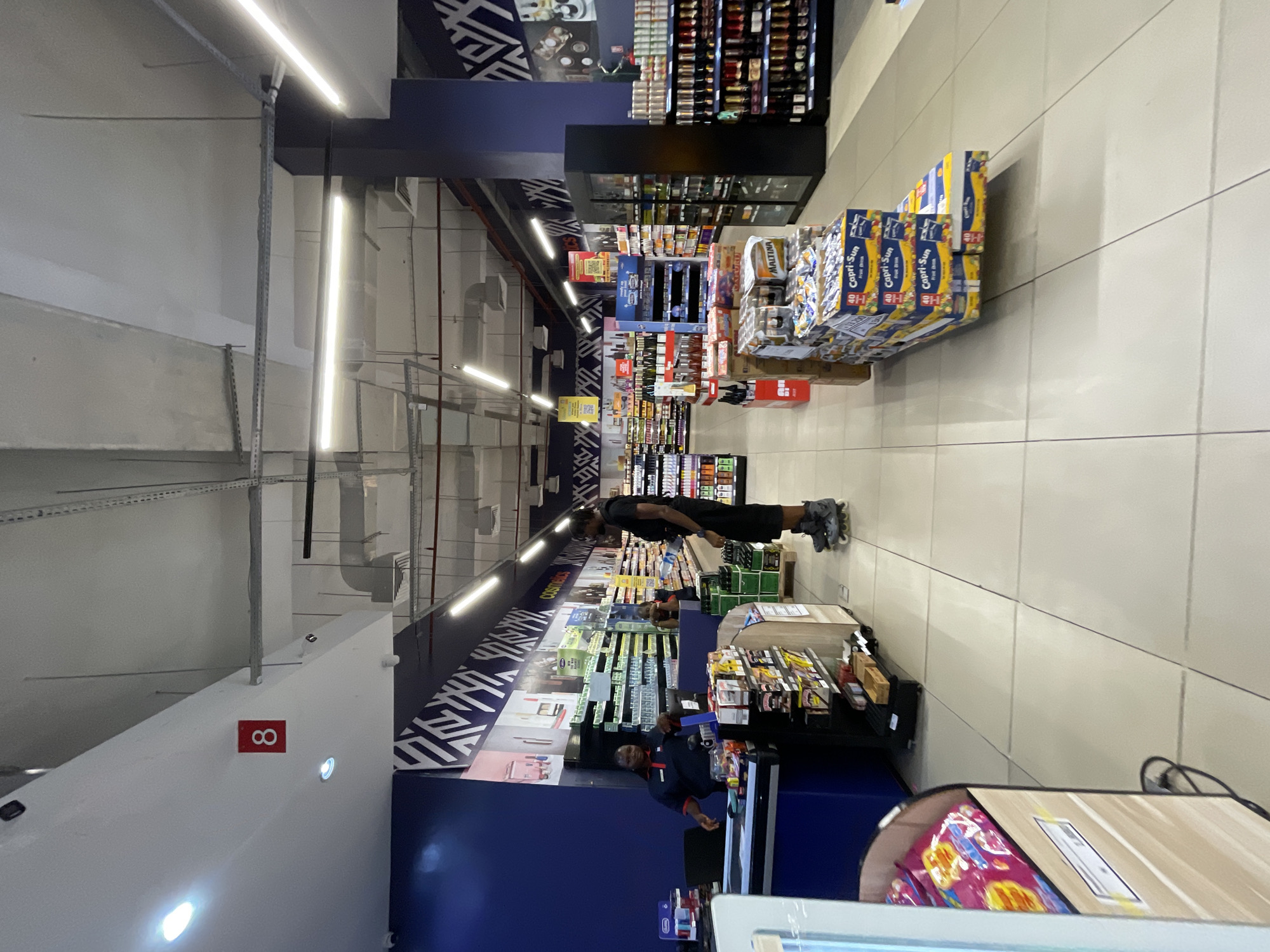 The image depicts the interior of a supermarket. In the foreground, to the left, a checkout counter is visible with a cashier wearing a blue uniform. A person on roller skates is standing near the checkout, facing away from the camera. Behind the checkout counter, shelves are stocked with various products. In the mid-ground, the aisles of the supermarket are lined with more shelves filled with groceries, including beverages and packaged goods. A large display of Capri-Sun juice boxes is prominent in the center of the image. The store features bright overhead lighting and a clean, tiled floor. The background shows more shelves and a blue wall with a decorative pattern. The overall impression is a well-organized retail environment. Based on the location provided, this is a supermarket in Moba, Nigeria.