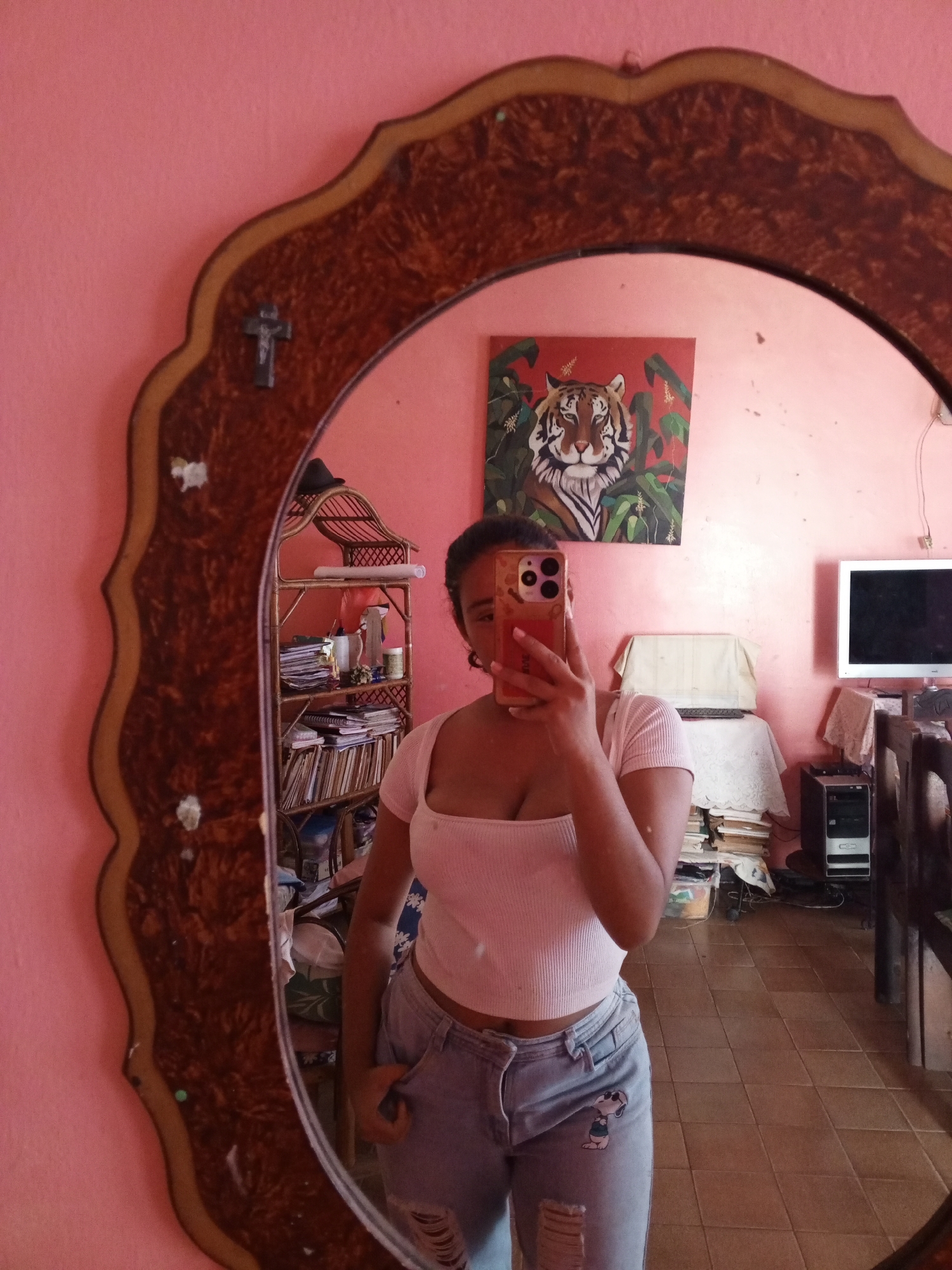 A person is taking a selfie in a mirror, which reflects a room with pink walls. The mirror has an ornate, dark brown frame with a lighter brown trim. In the reflection, a tiger painting hangs on the wall above a table covered with a white tablecloth, behind which is a television and computer tower. A bookshelf is visible to the left of the subject in the reflection. The person in the selfie is wearing a white crop top and light-wash denim jeans with a cartoon Snoopy patch. The person is holding a smartphone with a gold-colored case in front of their face, partially obscuring it. The location is Cúa, Venezuela.