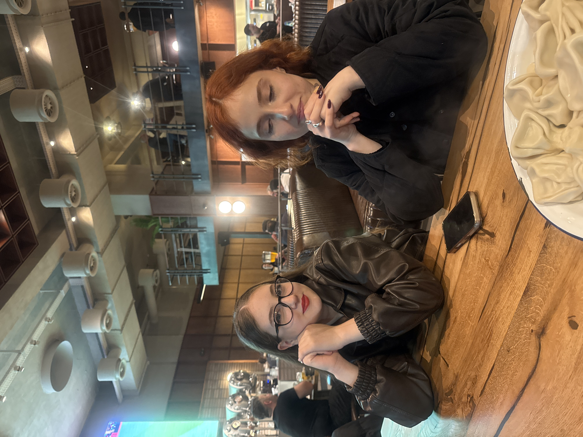 Two women are seated at a wooden table indoors. One woman has long auburn hair and is wearing a black shirt. The other woman has long brunette hair and is wearing glasses and a brown leather jacket. The woman with auburn hair has her hands clasped, and the woman with brunette hair has her chin resting on her hands. A plate of food, likely khinkali, is on the table. A black smartphone is also on the table. The location is Tbilisi, Georgia.
