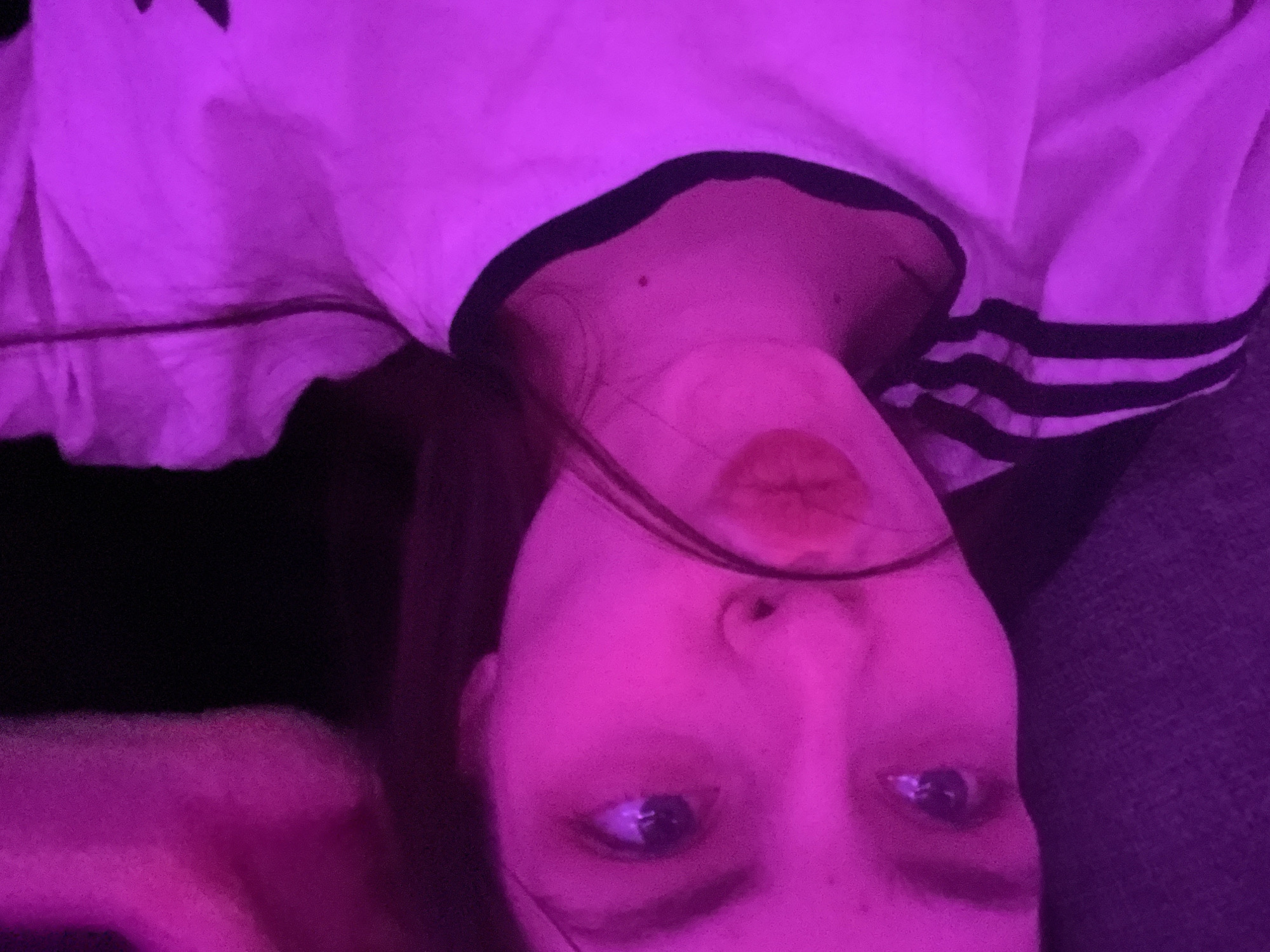 A selfie taken with the camera tilted upside down shows a person from the chest up in what appears to be a dark room lit by purple light. The person has light skin, visible eyes and lips, and some hair falling in front of their face. The person is wearing a white top with a black collar and what seems to be black stripes on the shoulder area. Some of the top is bunched on the left side of the frame. In the lower left corner, there is some blurred fabric. The background is dark. There is no evidence to suggest this photo was taken in Saint Petersburg, Russia.