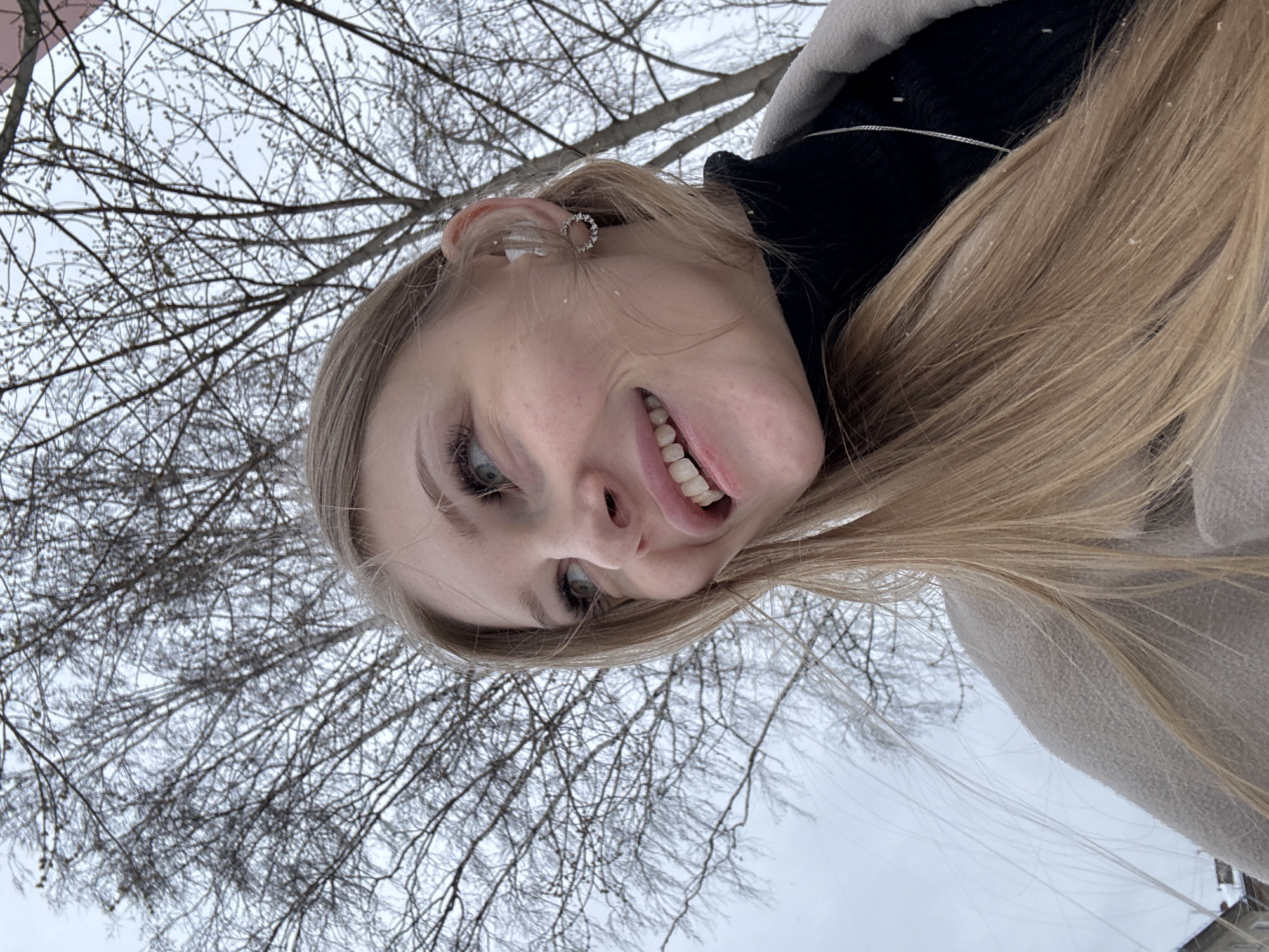 A woman is taking a selfie in what appears to be a cold, wintery environment. She has fair skin, blond hair, and is wearing an earring. In the background, there is a tree with bare branches against a pale sky. The presence of snow on her hair and the surrounding environment suggests that it has been snowing. The location is Perm, Russia.
