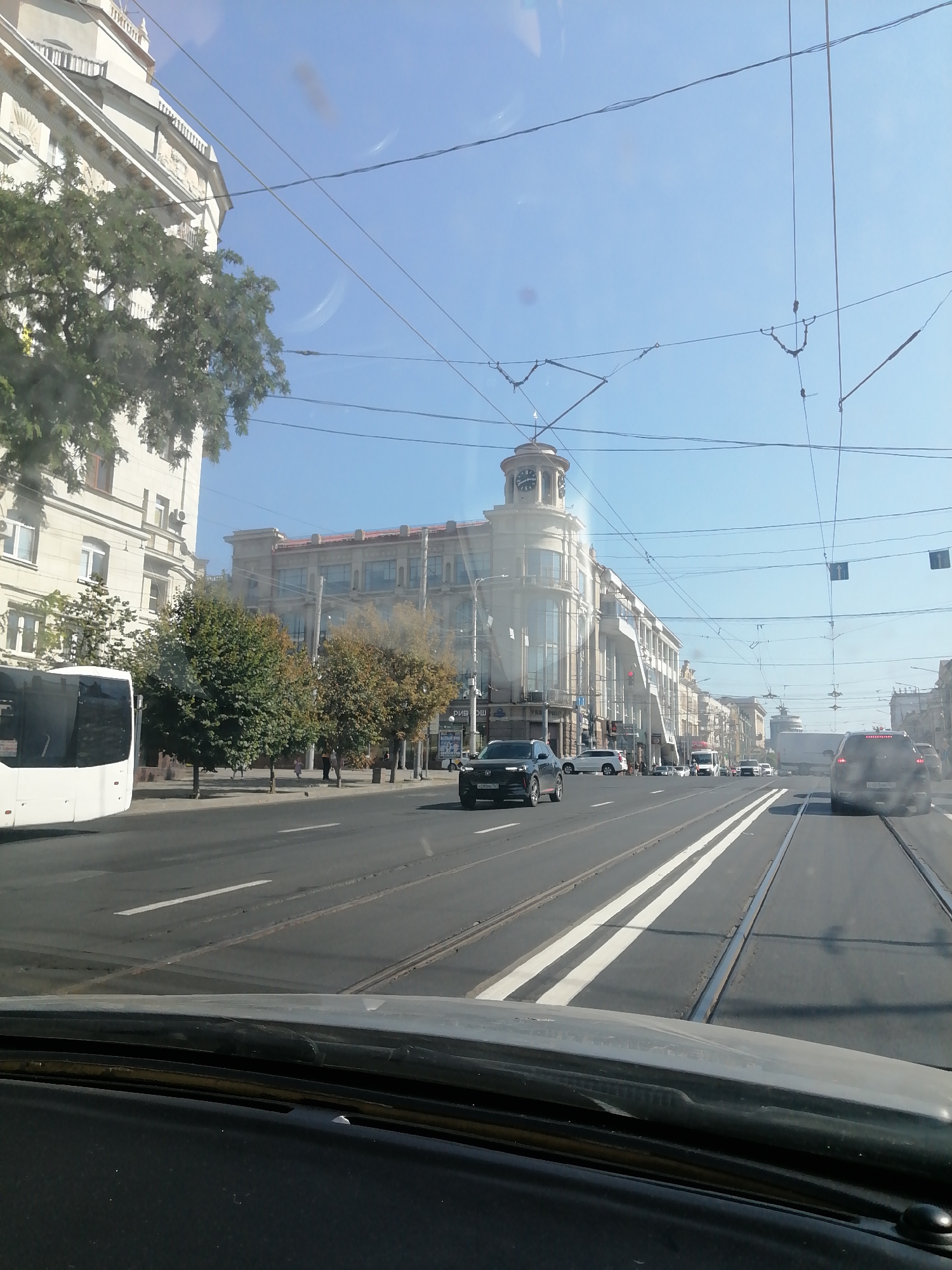 The image depicts a street scene in Rostov-on-Don, Russia, likely from inside a vehicle. In the foreground, the dashboard and windshield of a car are visible, suggesting the photo was taken from a driver's perspective. The street features tram tracks and is lined with buildings and trees. Several cars are on the road, with a white bus on the left and a dark SUV on the right. Overhead, a network of wires, possibly for trams or power, spans the sky, which is a clear blue. One building on the left has a clock tower. The overall atmosphere suggests a bright, daytime scene in an urban environment.