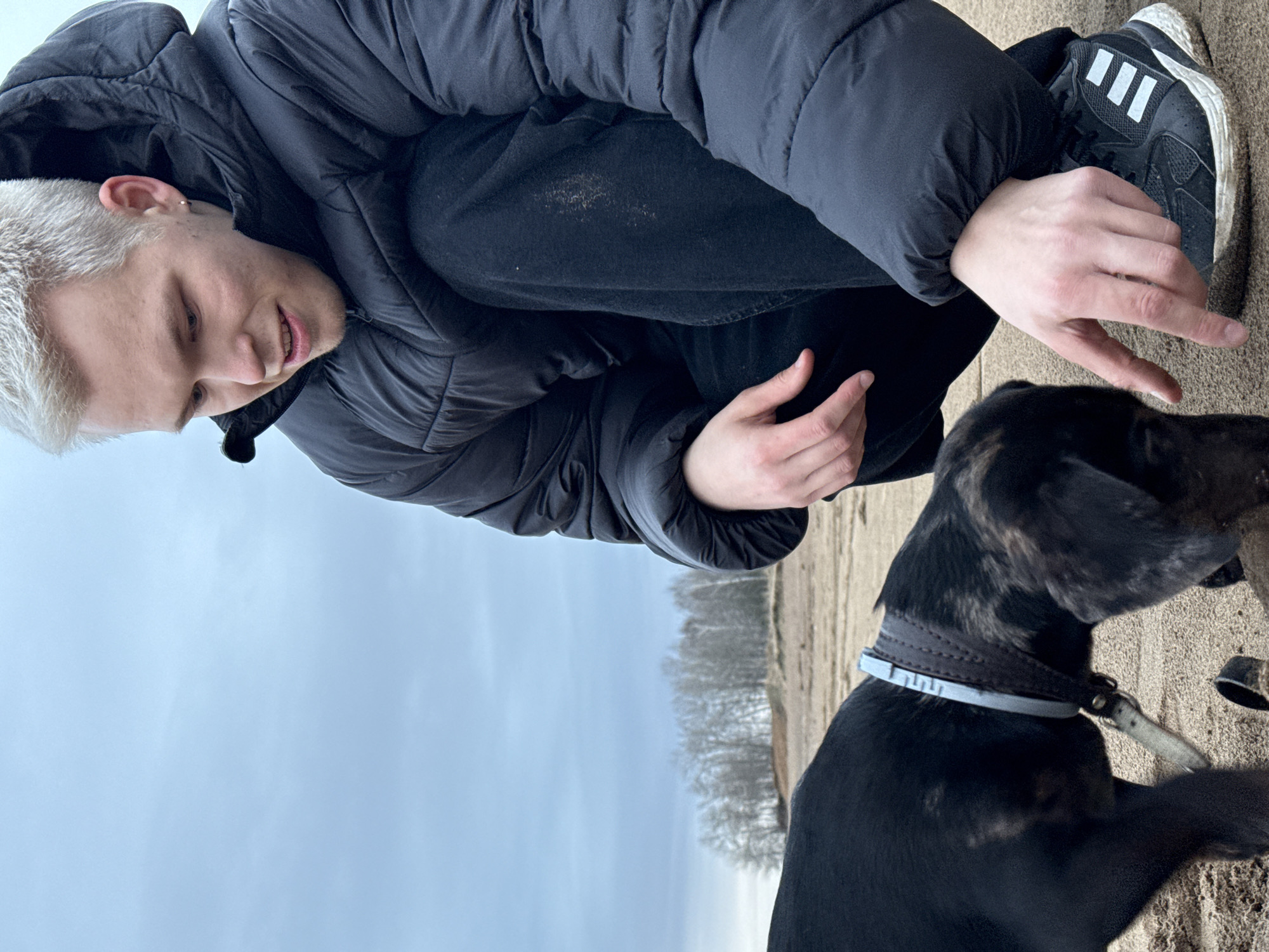 A young, light-skinned male with bleached blond hair is crouched low, extending a hand towards a black and brindle dog wearing a grey collar. The man is wearing a black puffer jacket, dark pants, and black athletic shoes with white stripes. The scene is set outdoors on a sandy or earthy ground, likely a park or natural area. In the background is a blurred view of trees and a cloudy sky, indicating possible overcast weather. The location is within the city of Chaikovsky, Russia.
