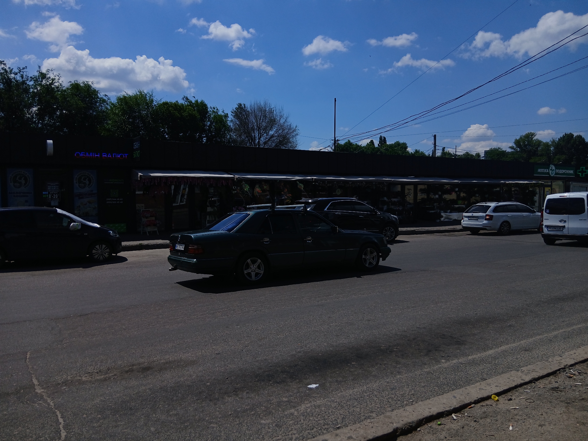 The image shows a street scene in Dnipro, Ukraine, under a bright blue sky with scattered white clouds. A dark-colored sedan is parked on the right side of the frame, and a white minivan is parked to its right. In the foreground, a dark-colored car is driving away from the viewer on the asphalt road. A row of shops with awnings is visible along the street. One shop has a neon sign that reads "ОБМІН ВАЛЮТ" (currency exchange). Another shop has a sign that reads "АПТЕКА ПОДОРОЖНИК" (Apteka Podorozhnik), which is a pharmacy. The road appears to be in fair condition with some visible cracks and patches. Trees and overhead power lines are visible in the background.