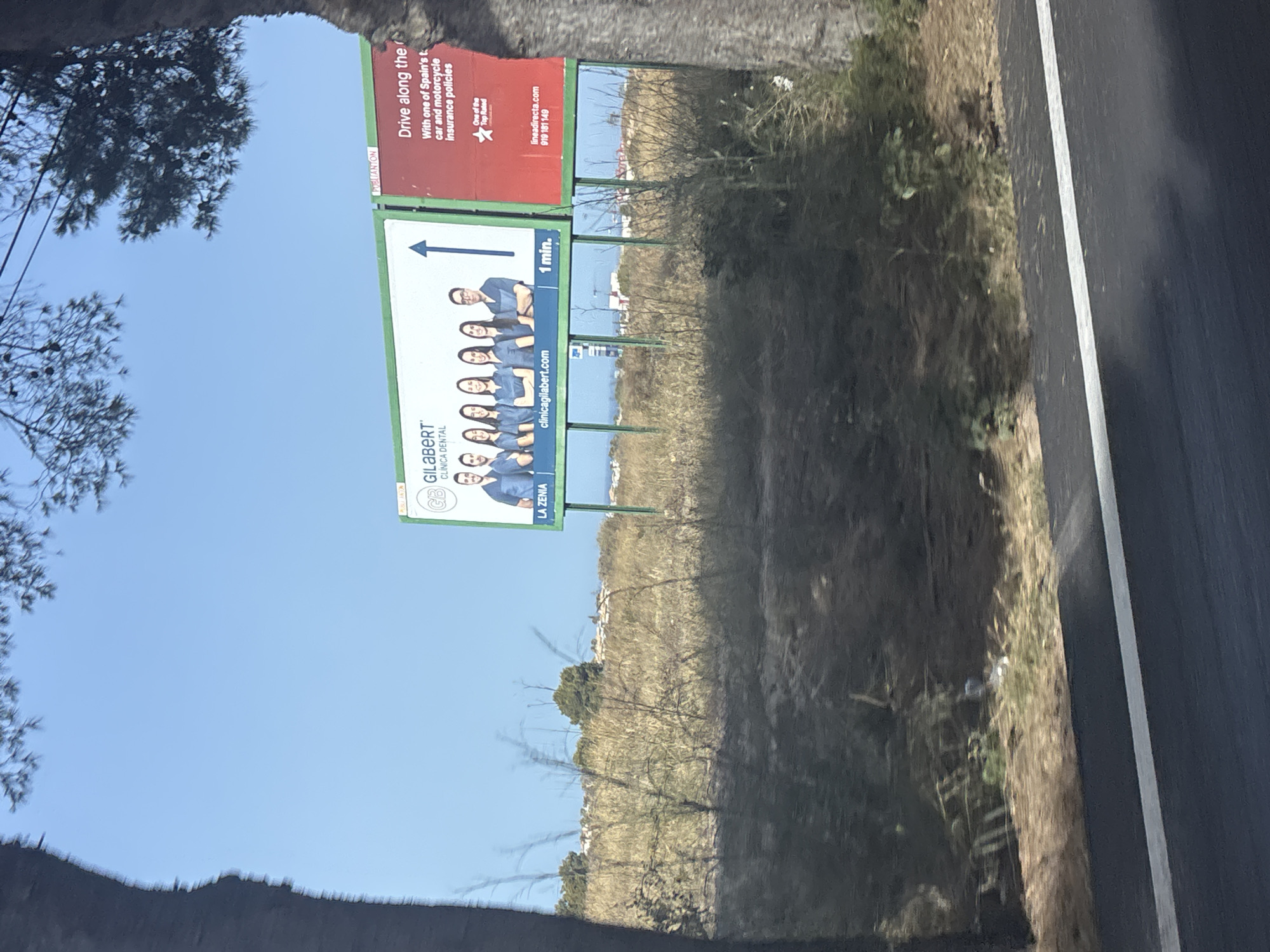 This media file is a photograph taken from a vehicle, likely while driving, as evidenced by the blurred foreground and the roadside setting. The image features two billboards along the side of a road.
The billboard on the right, which is red, advertises insurance policies for cars and motorcycles, stating "Drive along the... With one of Spain's t car and motorcycle Insurance policies". It also includes "lineadirecta.com" and a phone number, along with a mention of "One of the Top Rated".
The billboard on the left, which is white and green, advertises a dental clinic, "GILABERT CLÍNICA DENTAL", with the location "LA ZENIA" and the website "clinicagilabert.com". This billboard also displays a row of smiling people, presumably dentists or staff, and an arrow pointing to the right with "1 min." next to it.
The scene is outdoors, under a clear blue sky, suggesting daytime and fair weather. The surrounding environment appears to be a somewhat dry, grassy area with sparse vegetation and trees. In the distance, some buildings can be seen on a hillside. The perspective suggests the photo was taken from a moving vehicle on a paved road, with the right edge of the road clearly visible.