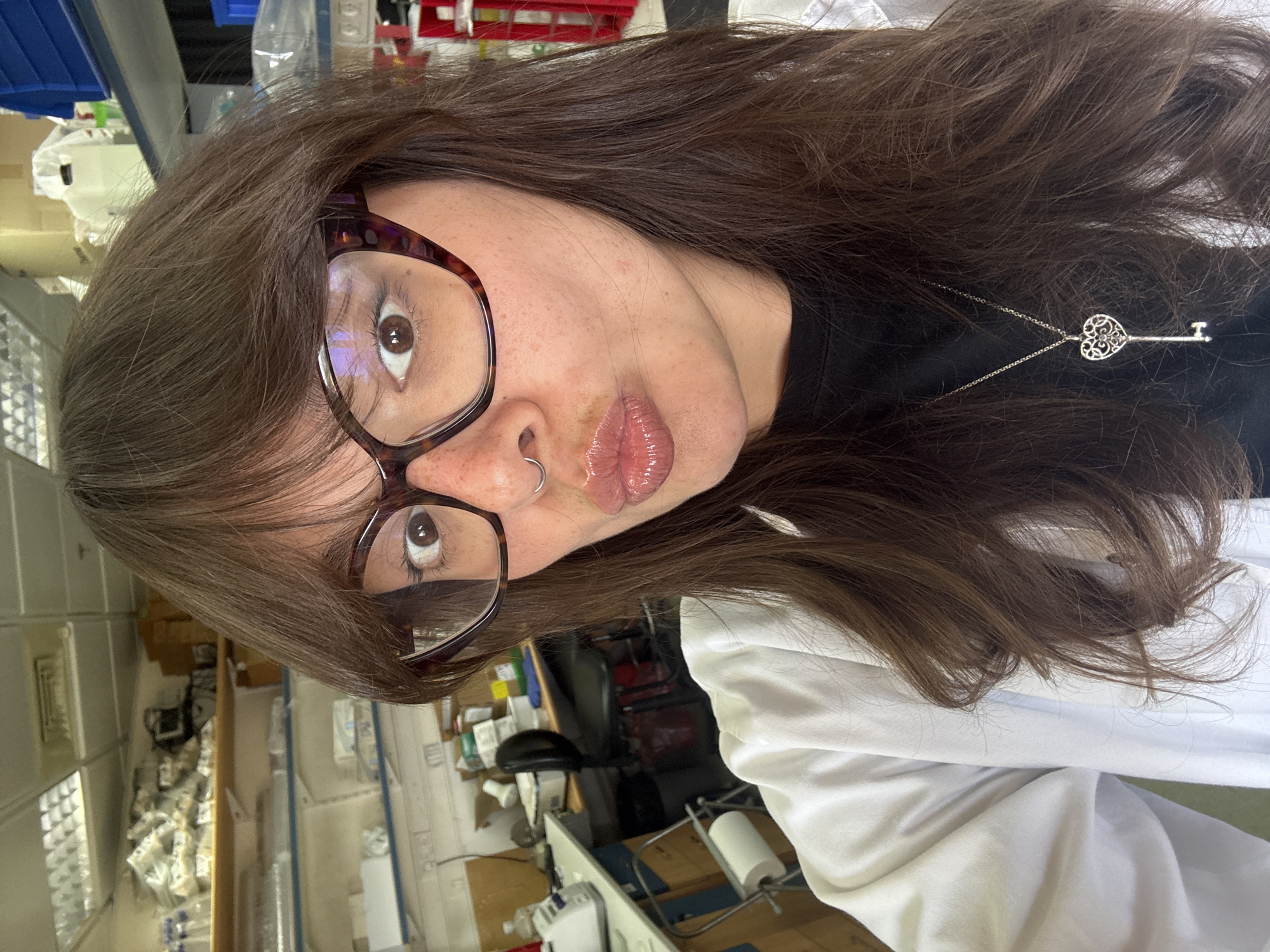 This is a portrait of a young person with long, dark brown hair, wearing large, tortoiseshell-patterned glasses. They are also wearing a silver necklace with a heart-shaped pendant and a black shirt. The person is in a room that appears to be a laboratory or medical setting, with shelves filled with medical supplies and equipment visible in the background. The lighting suggests it is daytime. The person is looking directly at the camera with their lips slightly pursed, and they have a nose ring. They are wearing a white lab coat, suggesting they are a healthcare professional or student. The overall tone of the image is casual and personal, likely a selfie taken in a workplace.