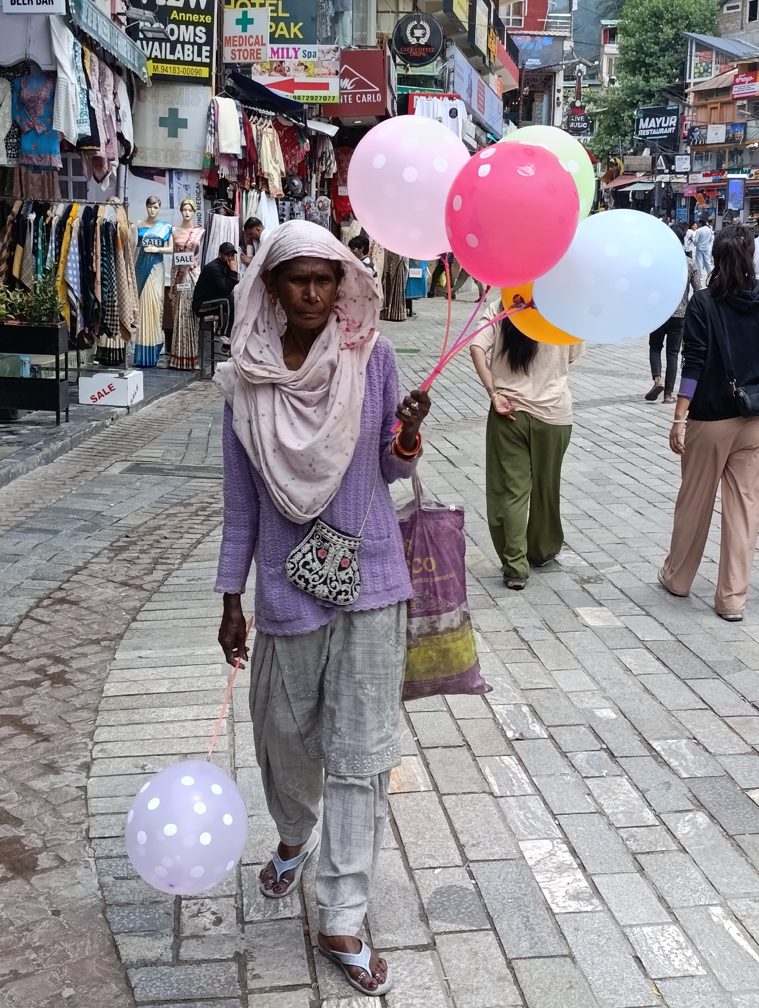 The image shows a street scene in Manali, India. A woman, appearing to be of elder age, walks towards the viewer holding a string attached to a light purple balloon with white polka dots. She is wearing a light purple sweater and a light pink headscarf, and gray patterned pants. She has a small embroidered purse hanging from her left shoulder. Behind her, a group of people are visible, including a woman holding a bunch of brightly colored balloons (pink, light green, and white), and other individuals walking along the street. The street is paved with stone tiles. Shops with signs in English and Hindi line the sides of the street, displaying clothing and other goods. One sign advertises "MAYUR RESTAURANT". The overall atmosphere appears to be that of a busy marketplace in a tourist town.