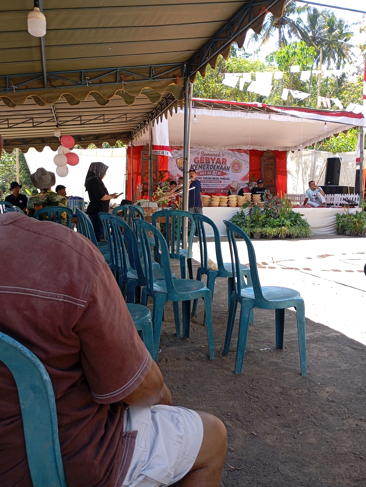 The image depicts an outdoor event in Ketepung, Indonesia, under a covered structure. In the foreground, a person wearing a brown t-shirt and white shorts is seated in a turquoise plastic chair, facing away from the camera. Multiple turquoise plastic chairs are arranged in rows. In the background, there is a stage set up with a banner that reads "Semarak GEBYAR KEMERDEKAAN HUT KE-81 RI". Several people are visible on and around the stage, some seated and some standing. A woman in a black headscarf and black clothing is standing near the front of the stage, holding a tablet. Decorative balloons, some pink and white, are suspended from the covered structure. Tropical trees and palm trees are visible behind the stage, suggesting a warm climate. The ground is covered in dirt and gravel.