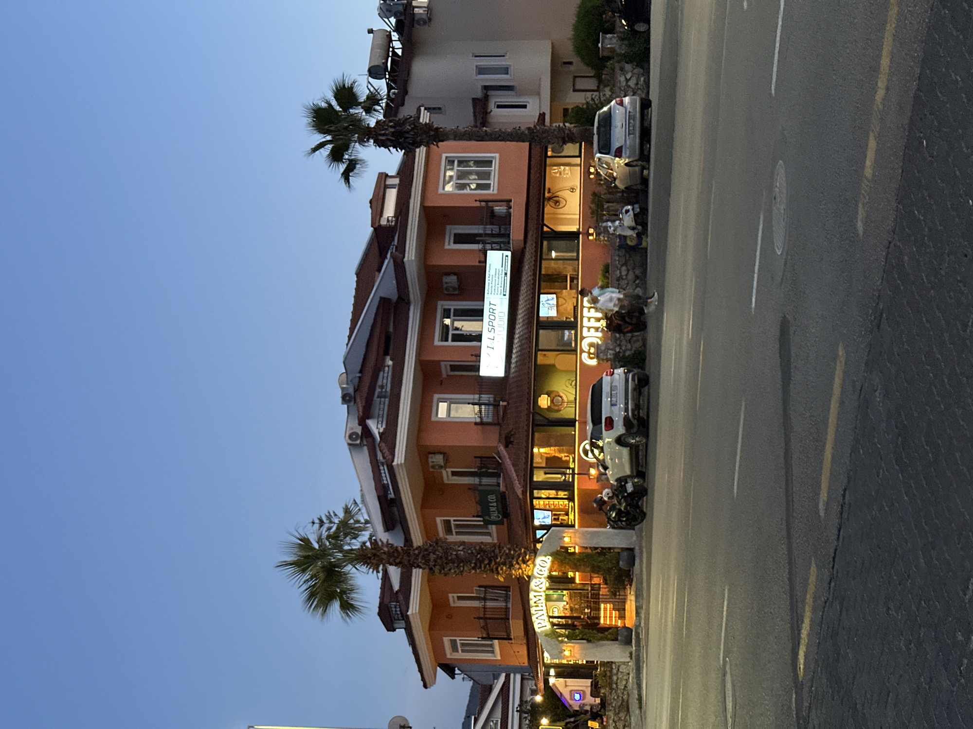 The image shows a street scene in Fethiye, Turkey at dusk. Several multi-story buildings line the street. Palm trees are adjacent to two buildings. The buildings appear to contain shops and restaurants, as indicated by signage such as "PALM & CO" and "COFFEE." Cars and motorcycles are parked along the side of the street. The street itself is paved and has lane markings. People can be seen near the entrances of the shops and restaurants. The sky is a gradient of blue, indicating the transition from day to night.