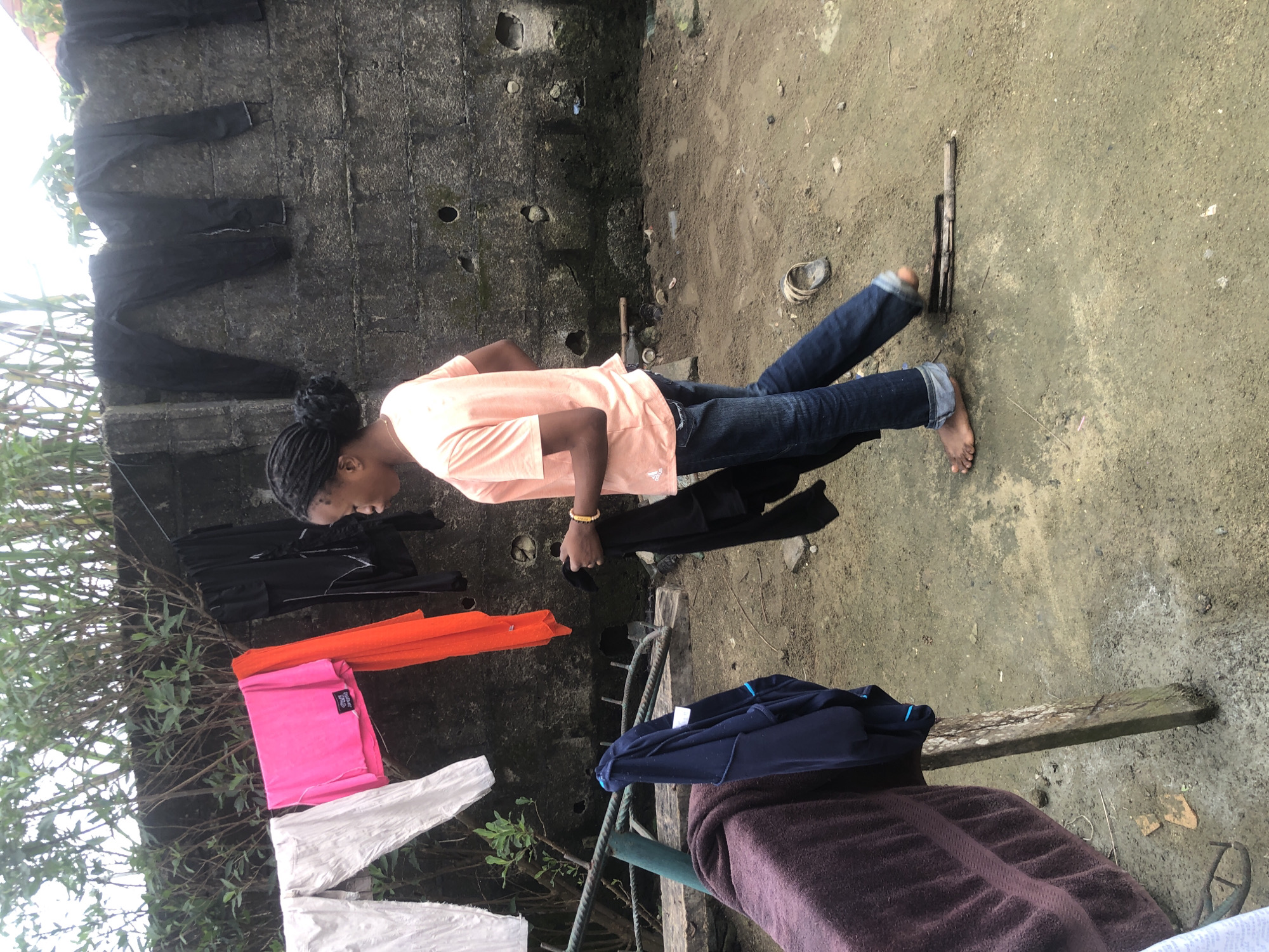 In Ado, Nigeria, a young person with dark braided hair is walking in an outdoor area with a concrete wall and lush green foliage in the background. The person is wearing a peach-colored t-shirt and blue jeans, and is holding a black piece of clothing. Laundry is hanging on a clothesline in front of the wall, including white pants, a pink top, an orange garment, and black pants. To the left, a purple towel and a dark blue jacket are draped over a wooden support structure. The ground is dirt and appears somewhat wet in places. A single sandal is visible on the ground. The overall scene depicts a casual domestic environment.