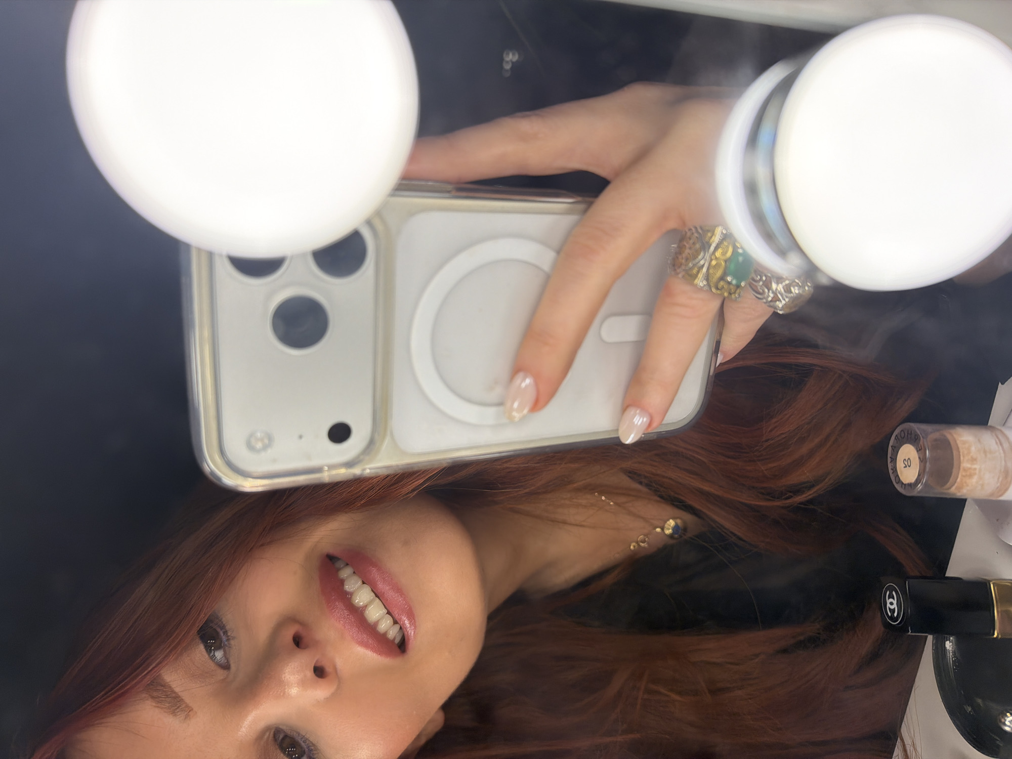 A selfie taken in a mirror shows a woman with long auburn hair and fair skin. She is holding a white smartphone with a ring light attachment, indicating she is using it to illuminate her face. Her teeth are visible in a smile, and her fingernails are manicured. She is wearing a ring on her right hand. To the right of the woman, makeup items are visible, including a Chanel product. It is not possible to determine from the image if it was taken in Paris, France.