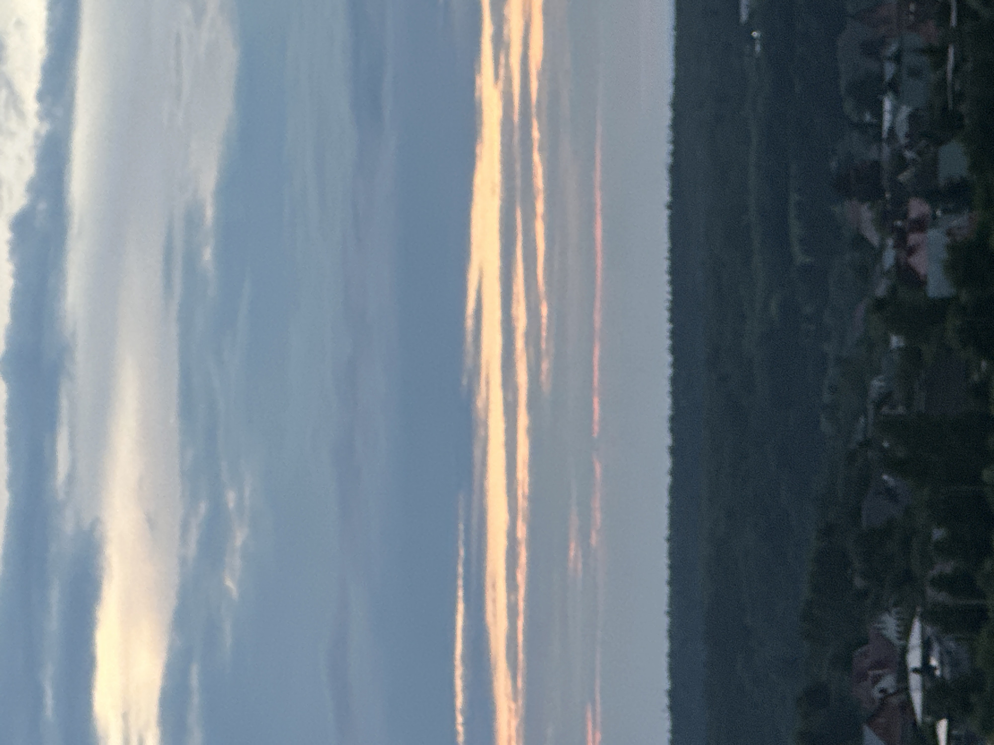 The image shows a distant view of a suburban area, likely in Podolsk, Russia, during twilight. The sky is filled with horizontal bands of clouds, with streaks of yellow and pink light indicating the setting sun. Below the sky, a dark, dense line of trees forms a horizon, suggesting a forested area or parkland. Beneath the trees, a scattering of houses with red and grey roofs are visible, indicating a residential neighborhood. The overall impression is of a quiet evening in a semi-rural setting.