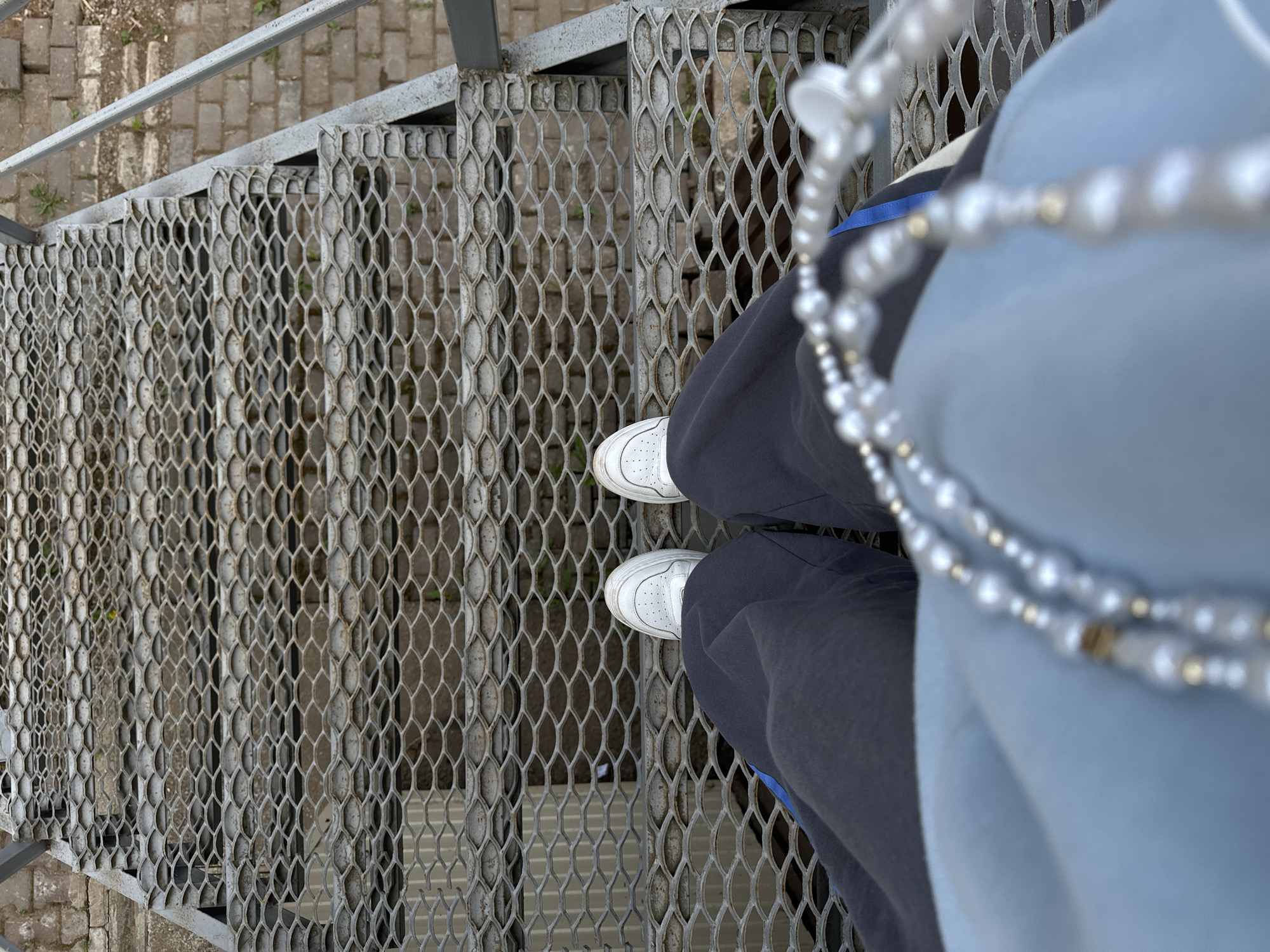 A low-angle shot depicts a person standing on a metal staircase, likely an industrial or utility structure. The staircase is made of a coarse, diamond-patterned metal grating. The person is wearing white sneakers, dark pants, and a light blue garment. A strand of white beads is draped across the person's torso. The environment surrounding the staircase consists of light gray brickwork and sparse vegetation, suggesting an outdoor setting. The city of Ilyinsky, Russia is not visually represented in this image.
